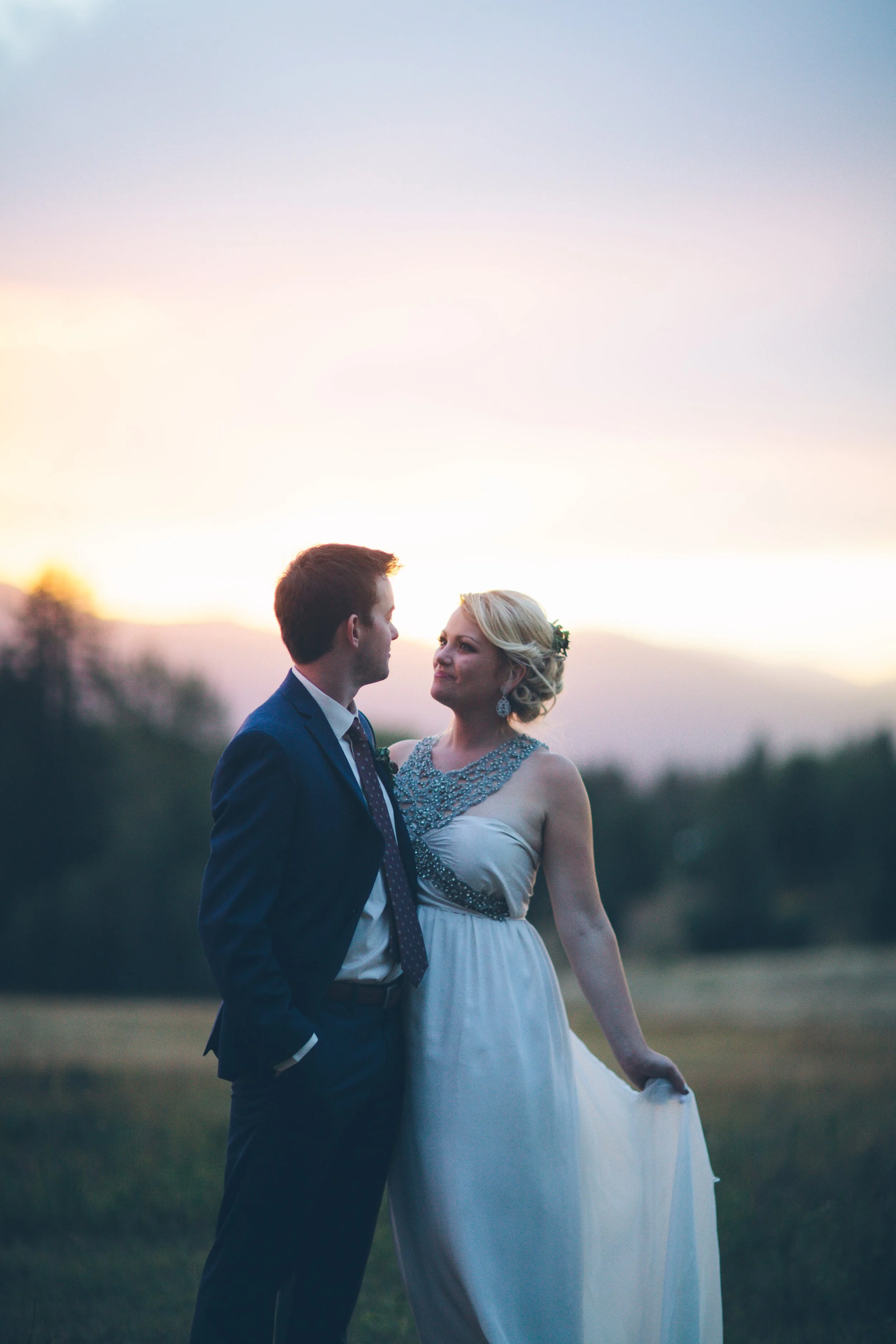 A bride and groom standing outdoors at sunset, gazing into each other's eyes with a soft glow in the background.