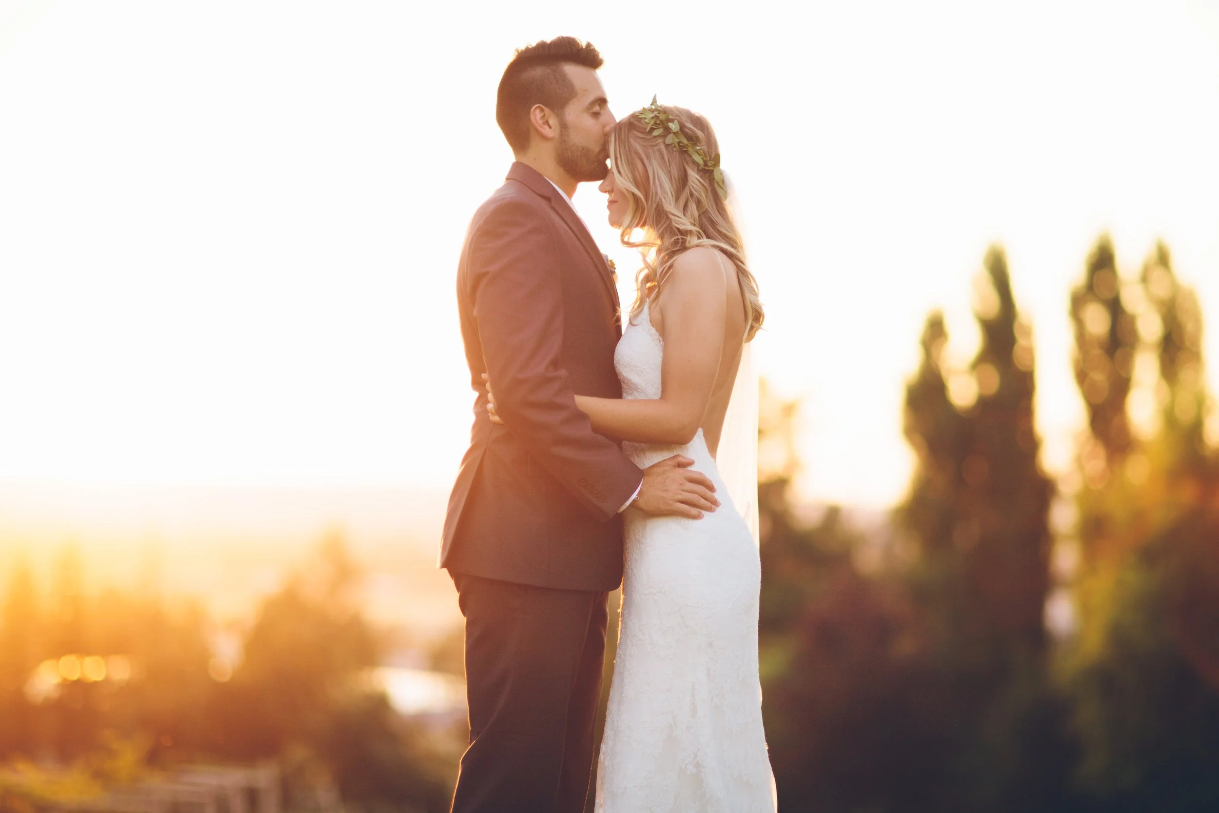A bride and groom standing close together during sunset, embracing and about to kiss, with trees blurred in the background.