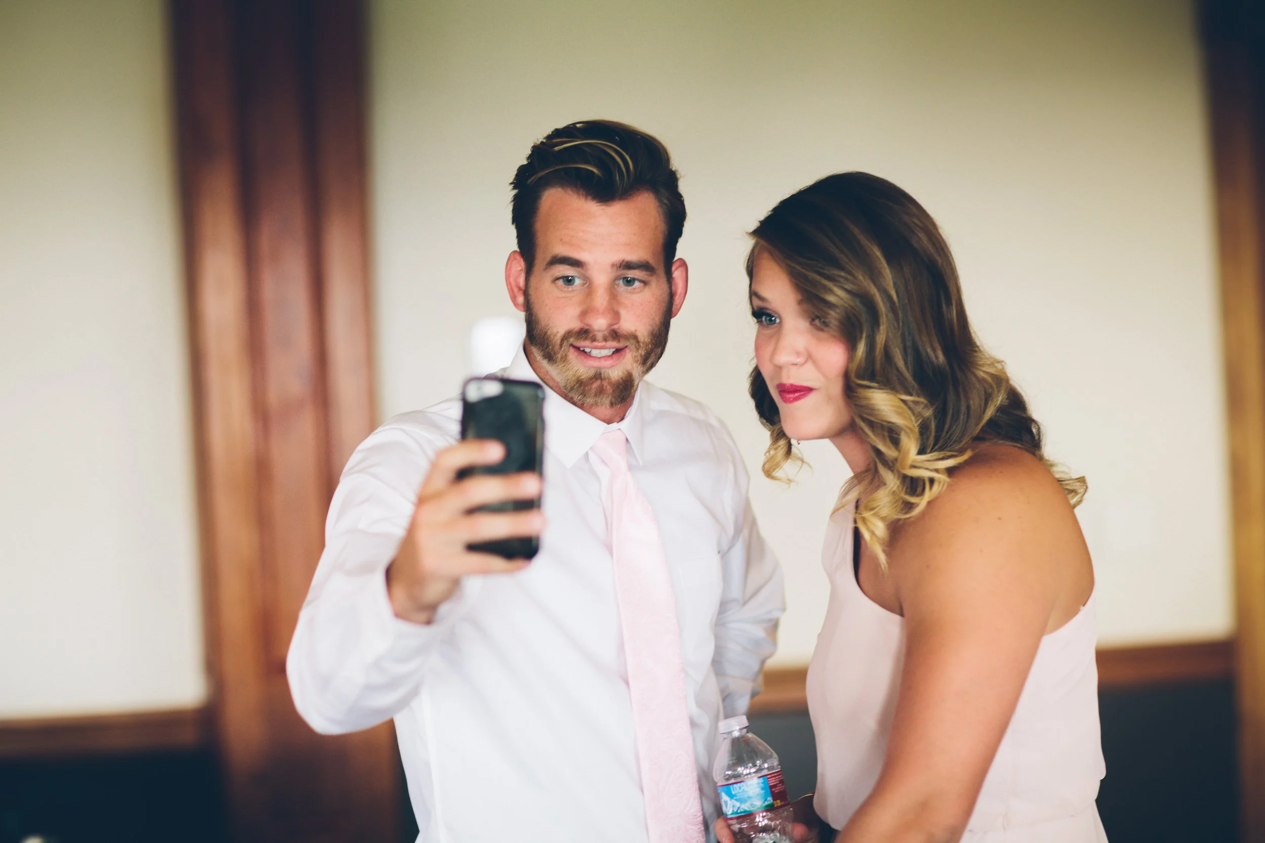 A man and woman taking a selfie together indoors, the man is holding a water bottle and the woman is wearing a sleeveless dress.