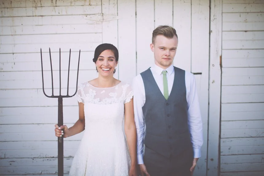 A woman in a wedding dress holding a pitchfork, standing next to a man in a vest and tie, both standing in front of a white wooden wall.