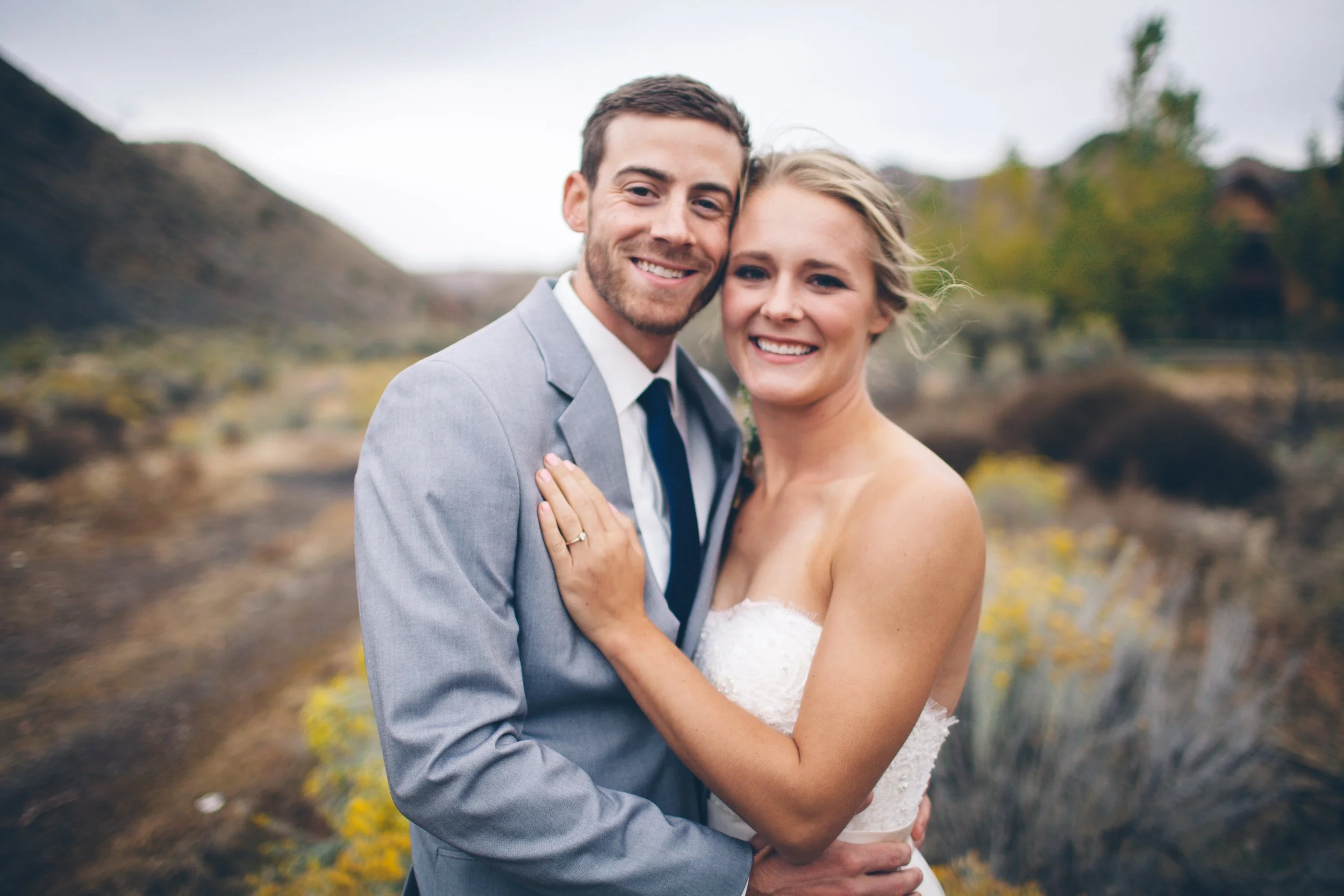 A smiling couple, the man in a gray suit and the woman in a white wedding dress, embracing outdoors in a natural setting with hills and trees in the background.