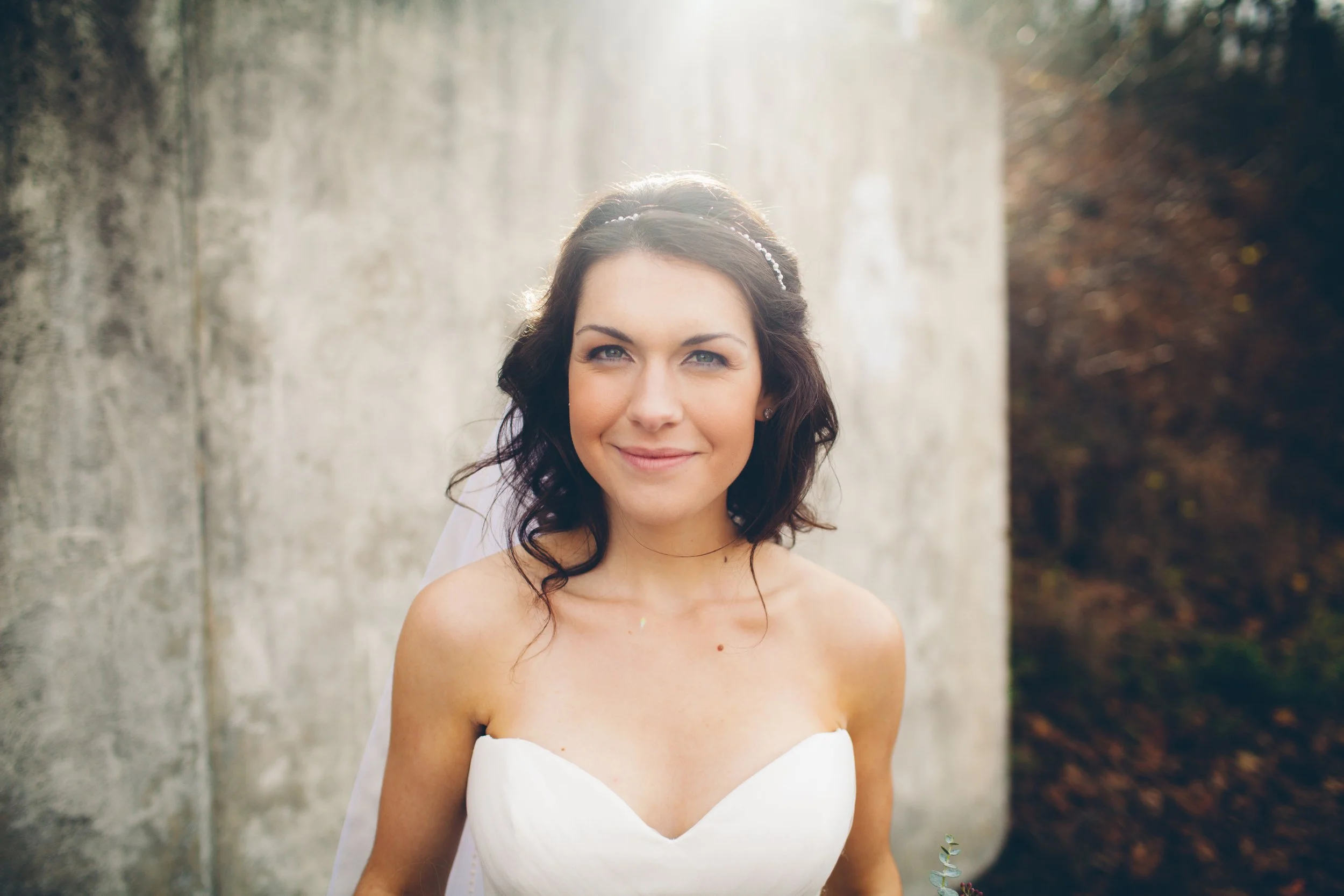 A woman in a white strapless wedding dress outdoors with a stone wall behind her and sunlight shining on her hair.