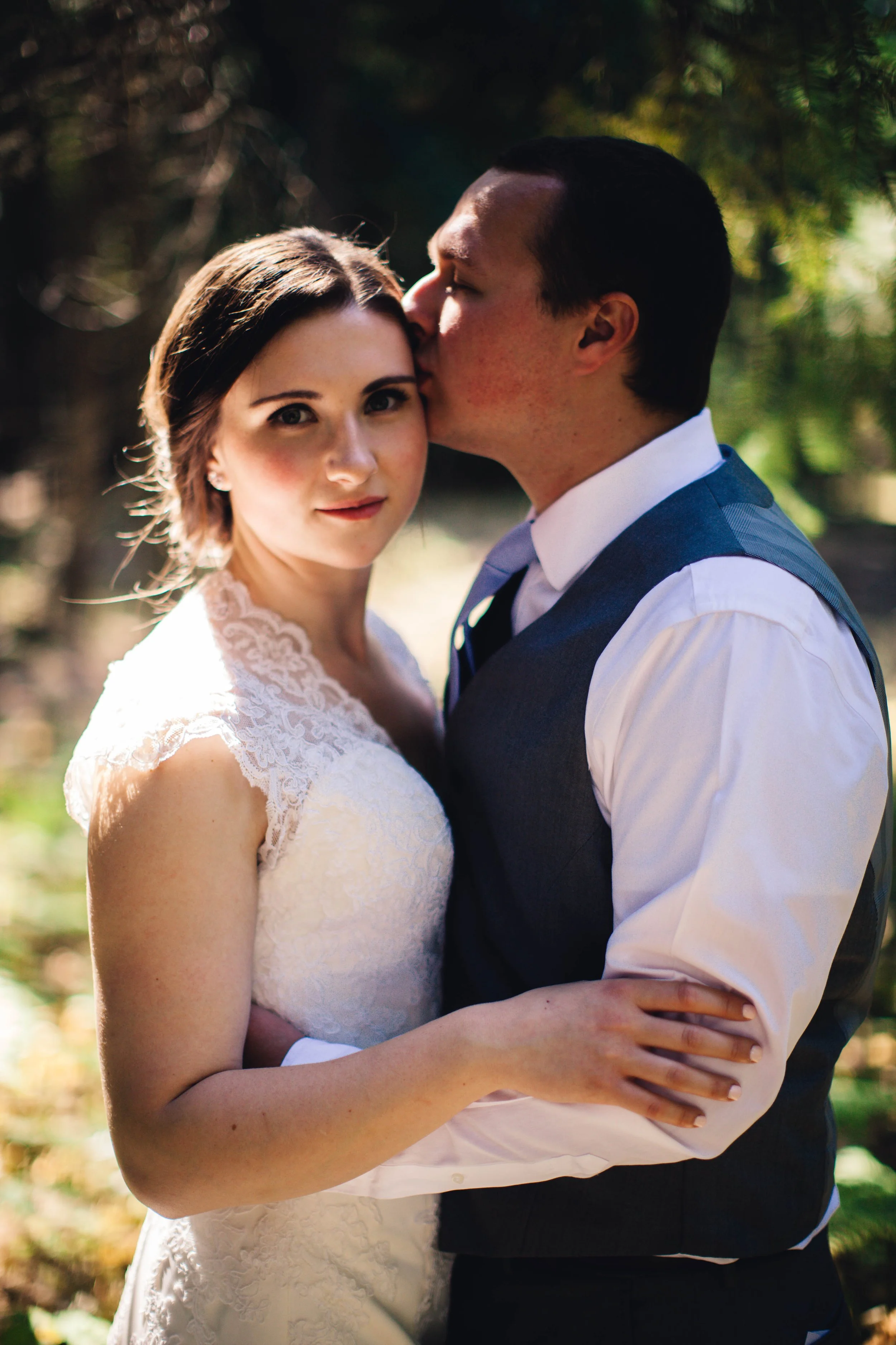 A bride and groom in wedding attire embracing outdoors, with the groom kissing the bride's temple, under a canopy of trees.