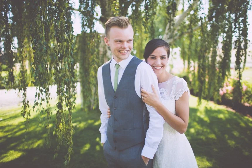 A happy couple in wedding attire smiling outdoors under trees.