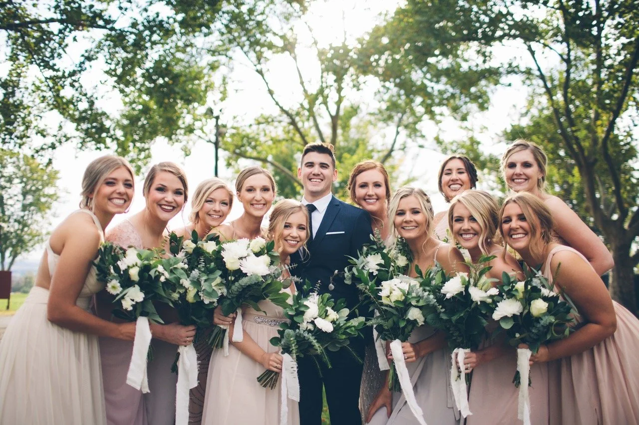 A group of women and a man in a wedding party outdoors, smiling and holding bouquets of white flowers, with trees in the background.