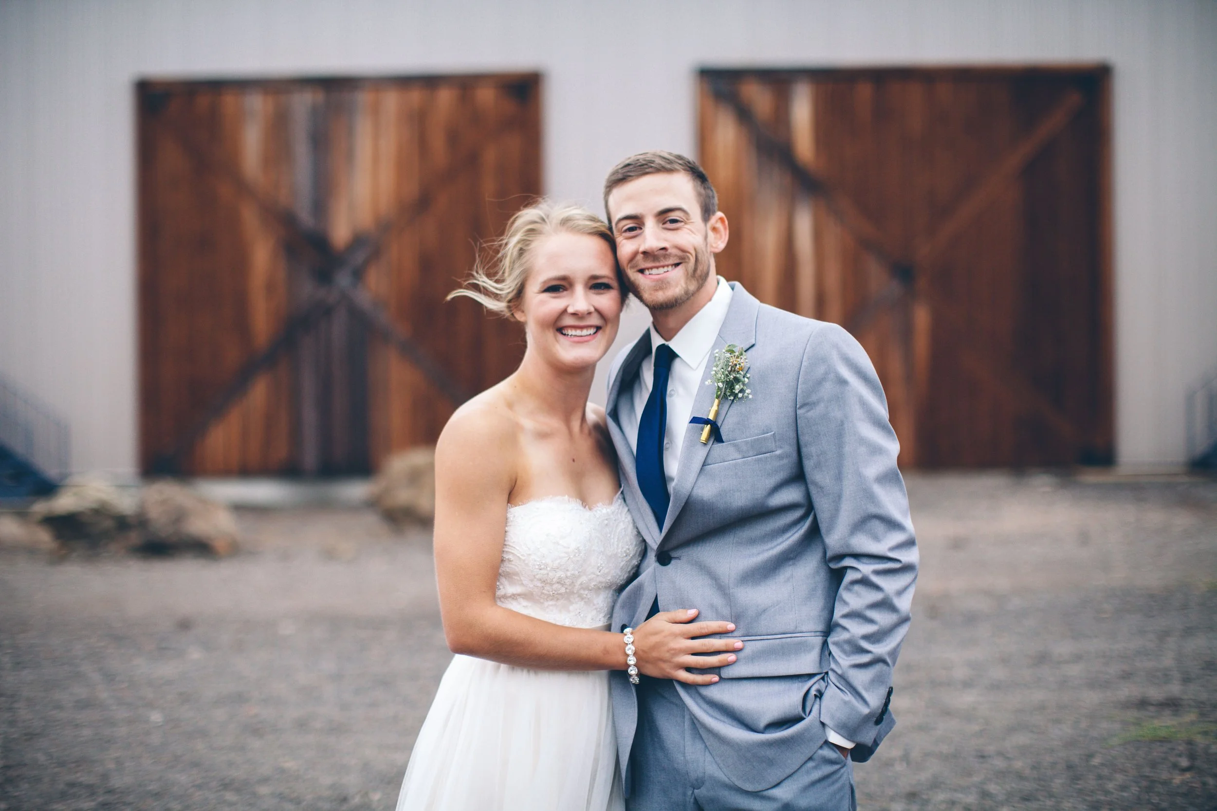 A smiling bride and groom standing close together outdoors, posing for a wedding photo in front of rustic wooden barn doors. The bride wears a strapless white wedding dress and the groom wears a light gray suit with a white shirt, navy tie, and bouto
