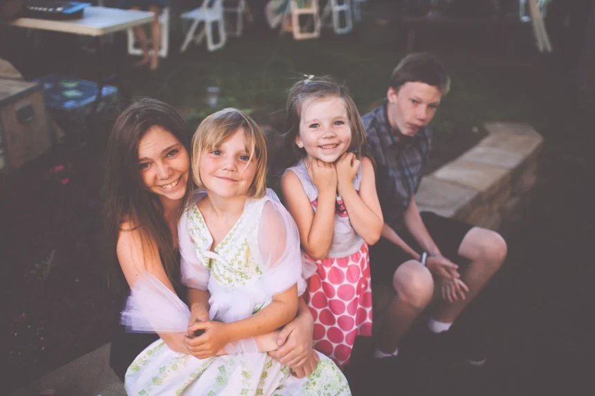 Four children sitting outdoors at an event, two girls smiling at the camera, a girl with arms crossed smiling, and a boy looking at the camera with a serious expression.