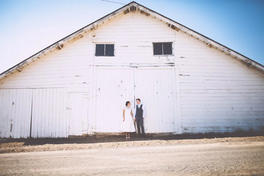 A man and woman holding hands in front of a large white barn with a gabled roof, set against a clear blue sky.