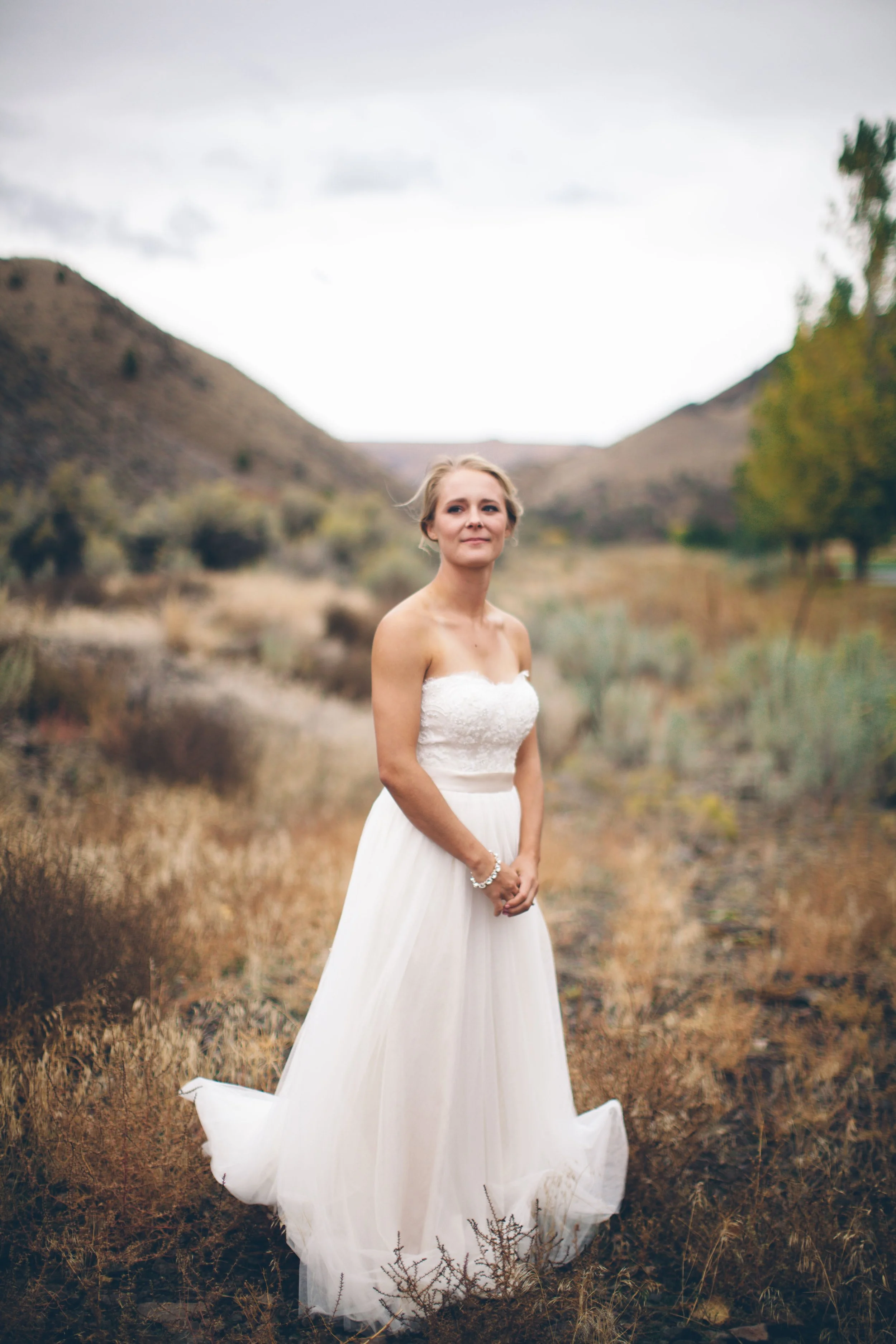 A woman wearing a white wedding dress standing outdoors in a natural, mountainous landscape with dry grass and shrubs.
