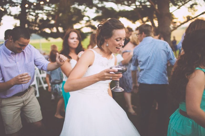 Woman in a wedding dress dancing at an outdoor celebration with other guests, holding a wine glass.