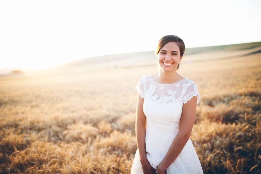 Young woman in a white lace dress smiling in a golden field at sunset.