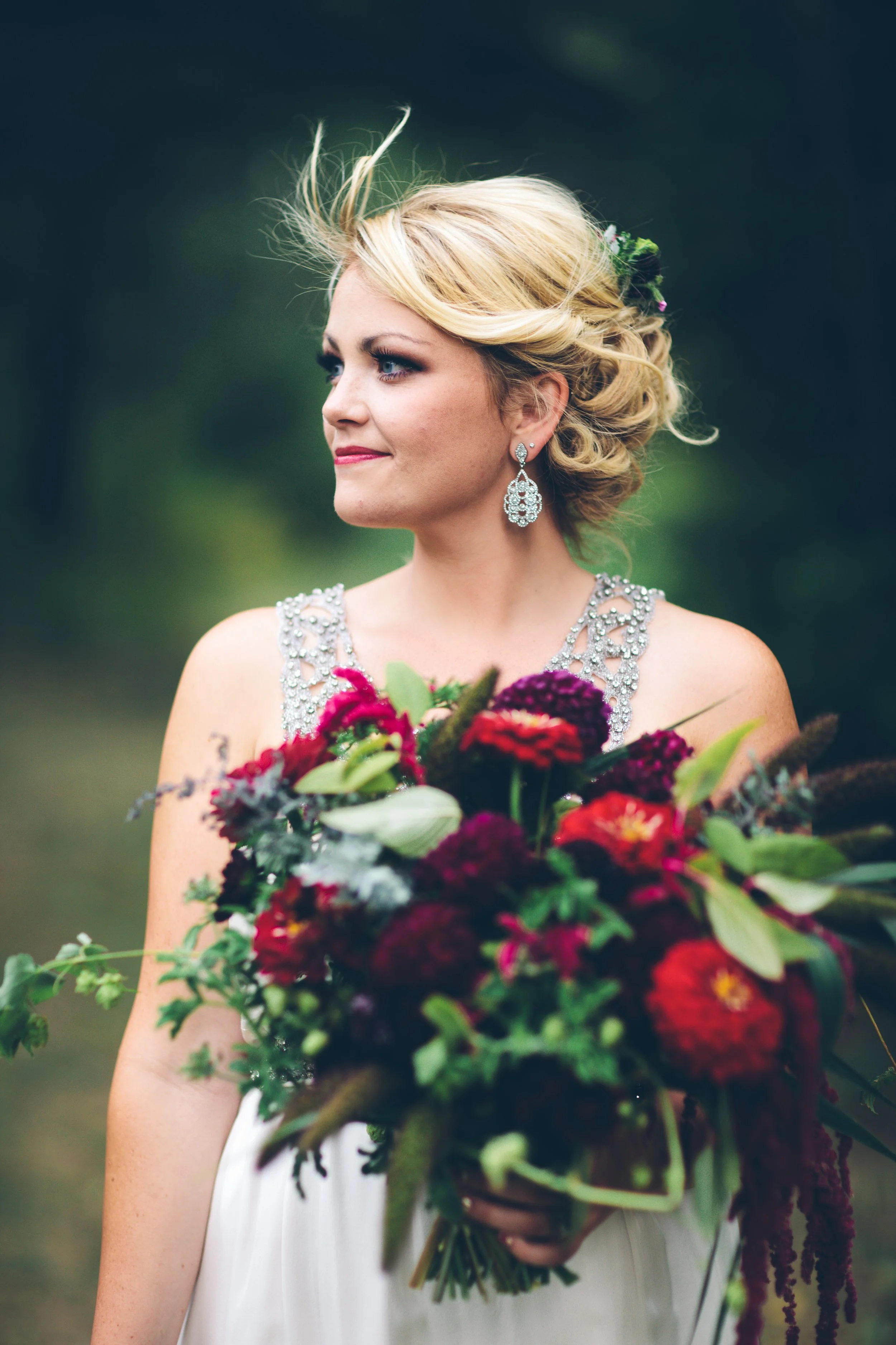 A bride with blonde hair in an updo, wearing earrings and a decorated dress, holding a large bouquet of red and purple flowers outdoors.