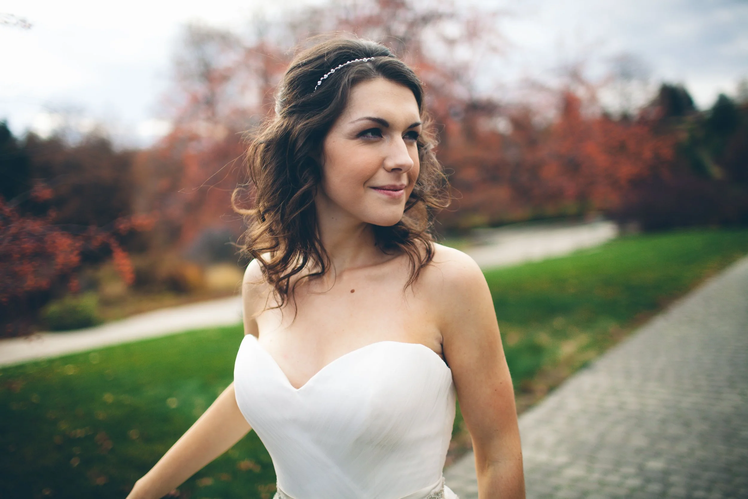 A bride in a white strapless wedding dress outdoors with autumn trees in the background.