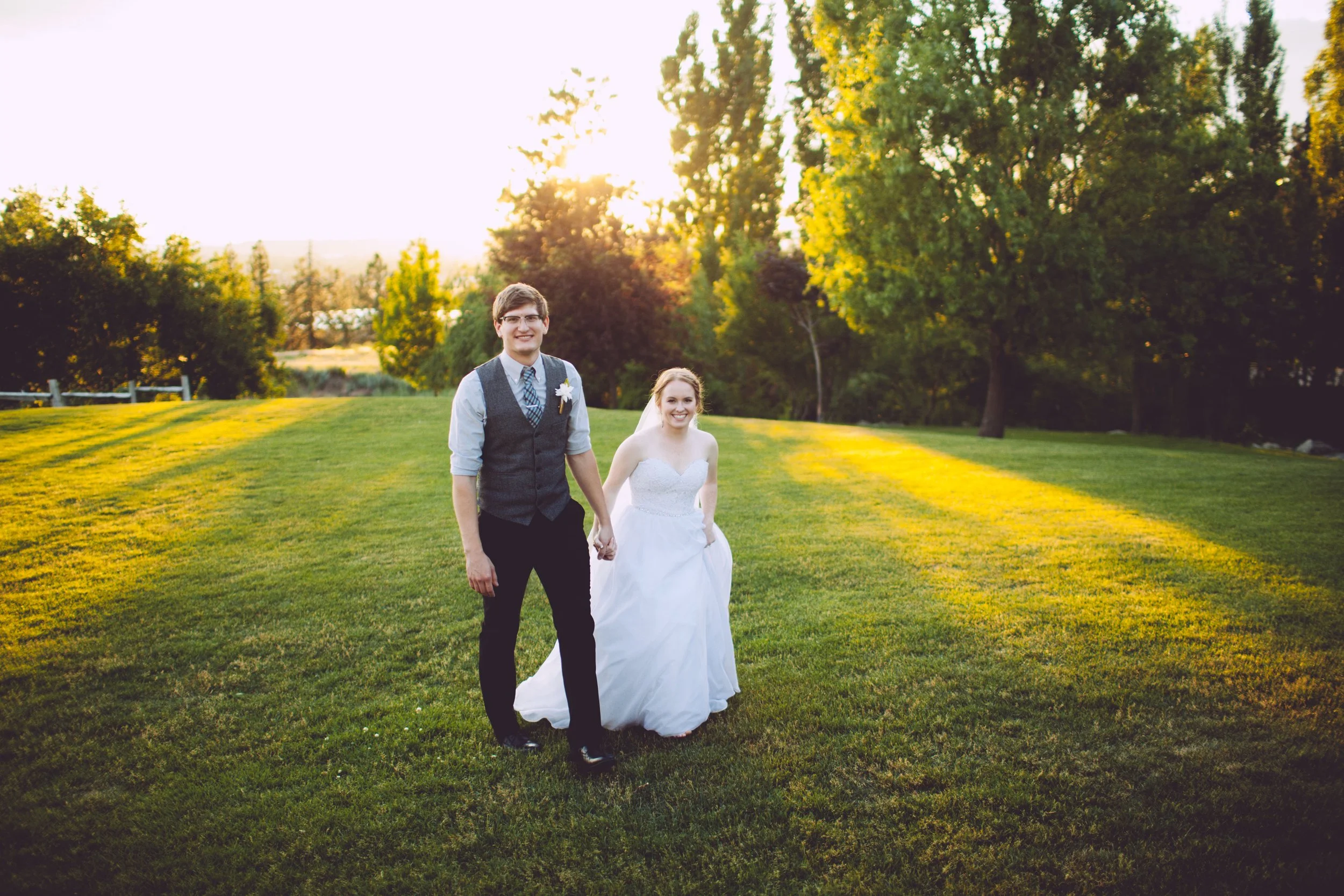 A newlywed couple holding hands and walking across a grassy field during sunset, surrounded by trees.
