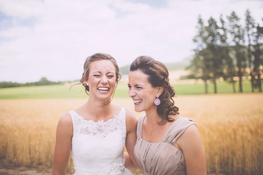 Two women laughing and smiling outdoors in a field with trees and sky in the background, one in a white dress and the other in a beige dress.