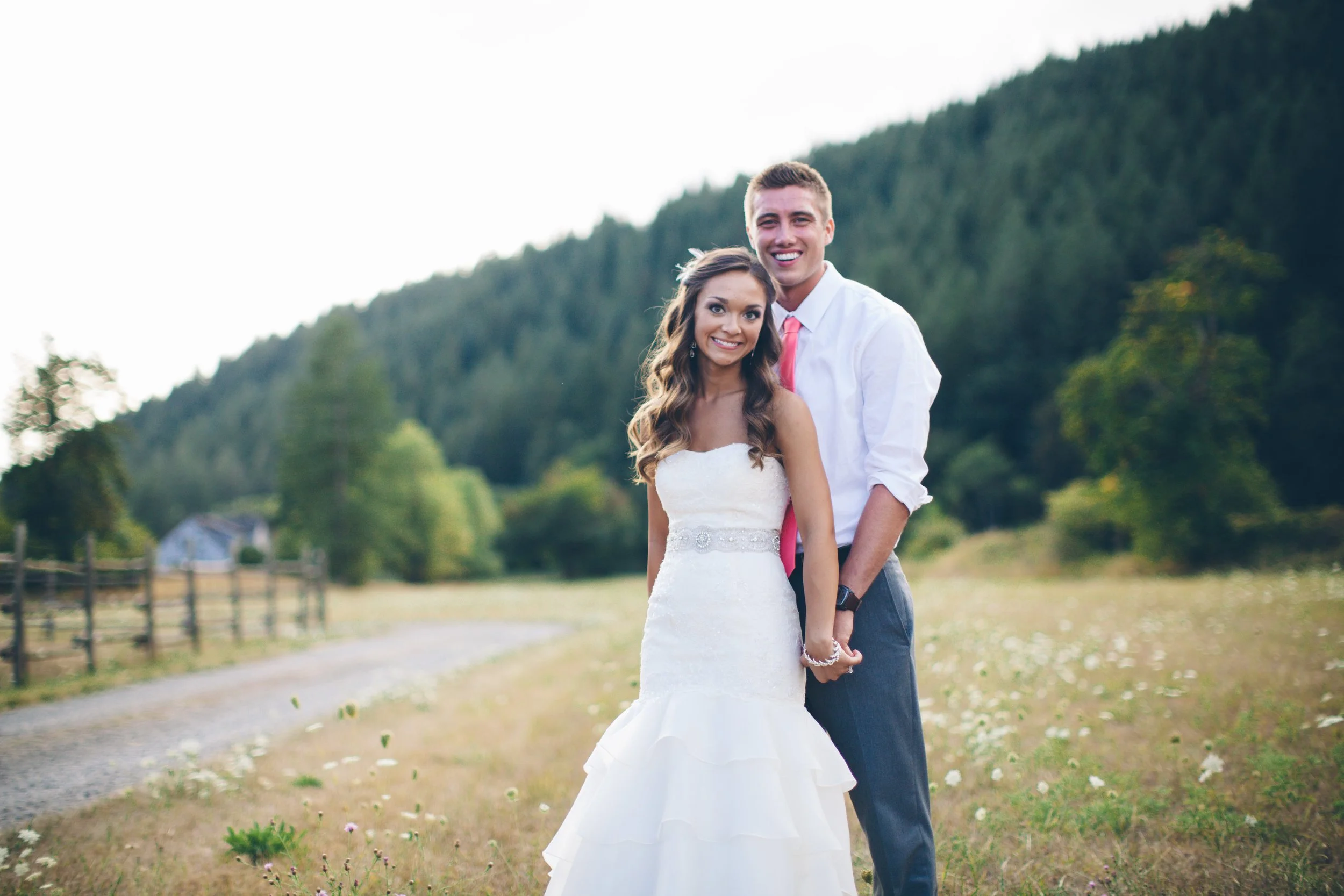A happy couple dressed in wedding attire standing outdoors in a grassy field with trees and mountains in the background.