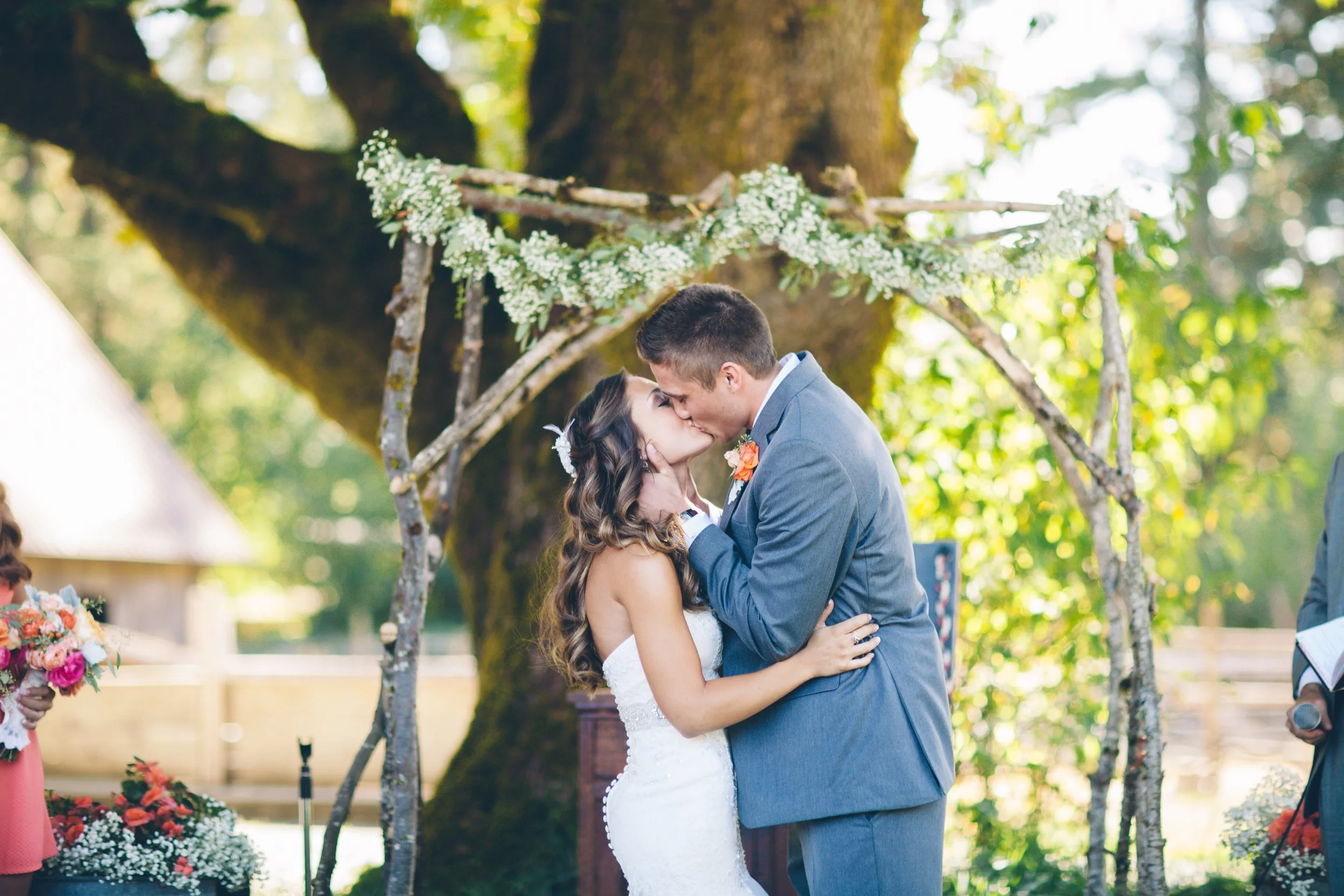 A bride and groom kissing under a rustic wedding arch in an outdoor setting with trees and sunlight.