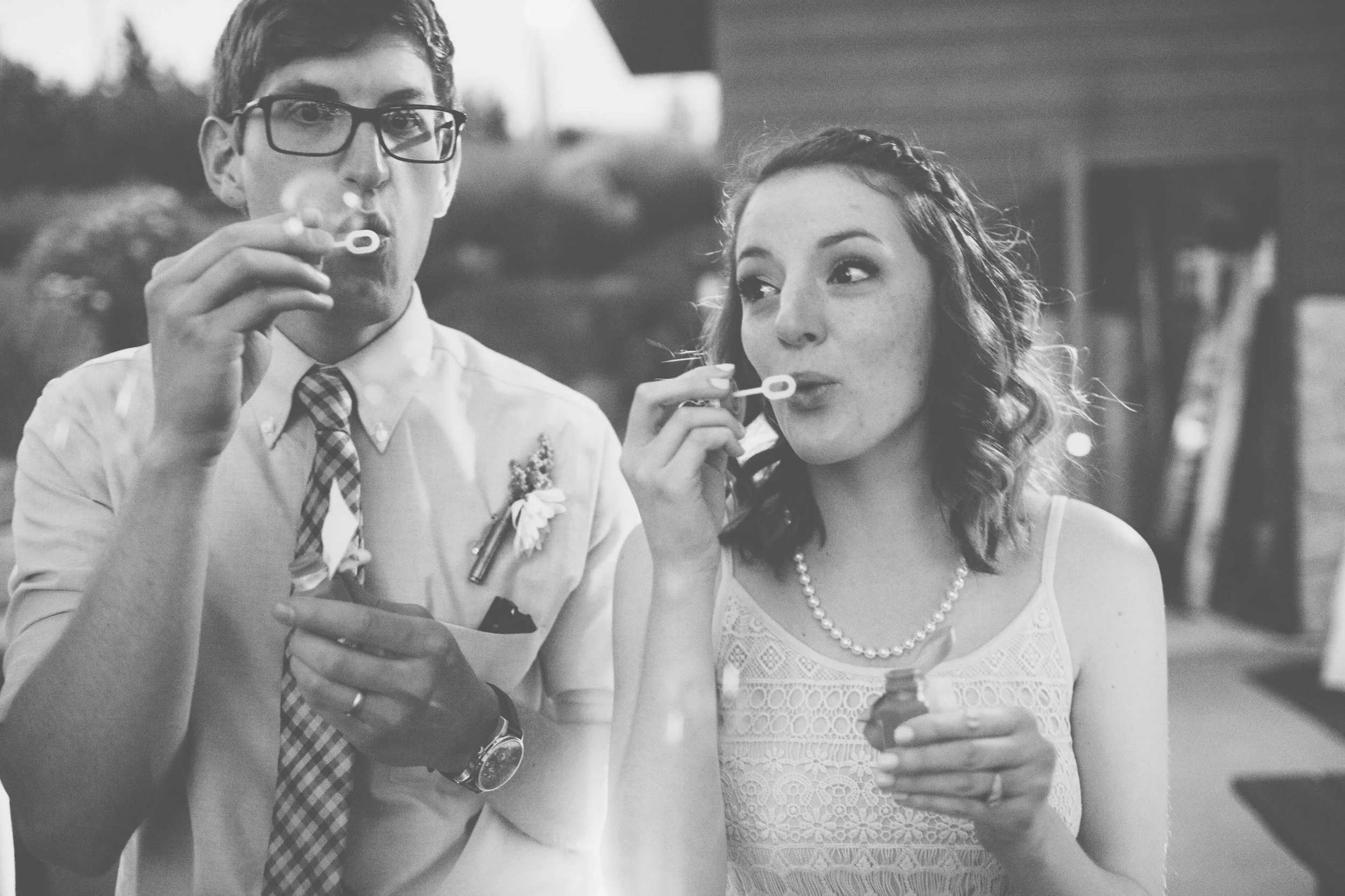 Black and white photo of a man and a woman blowing bubbles at an outdoor gathering, dressed in semi-formal attire.