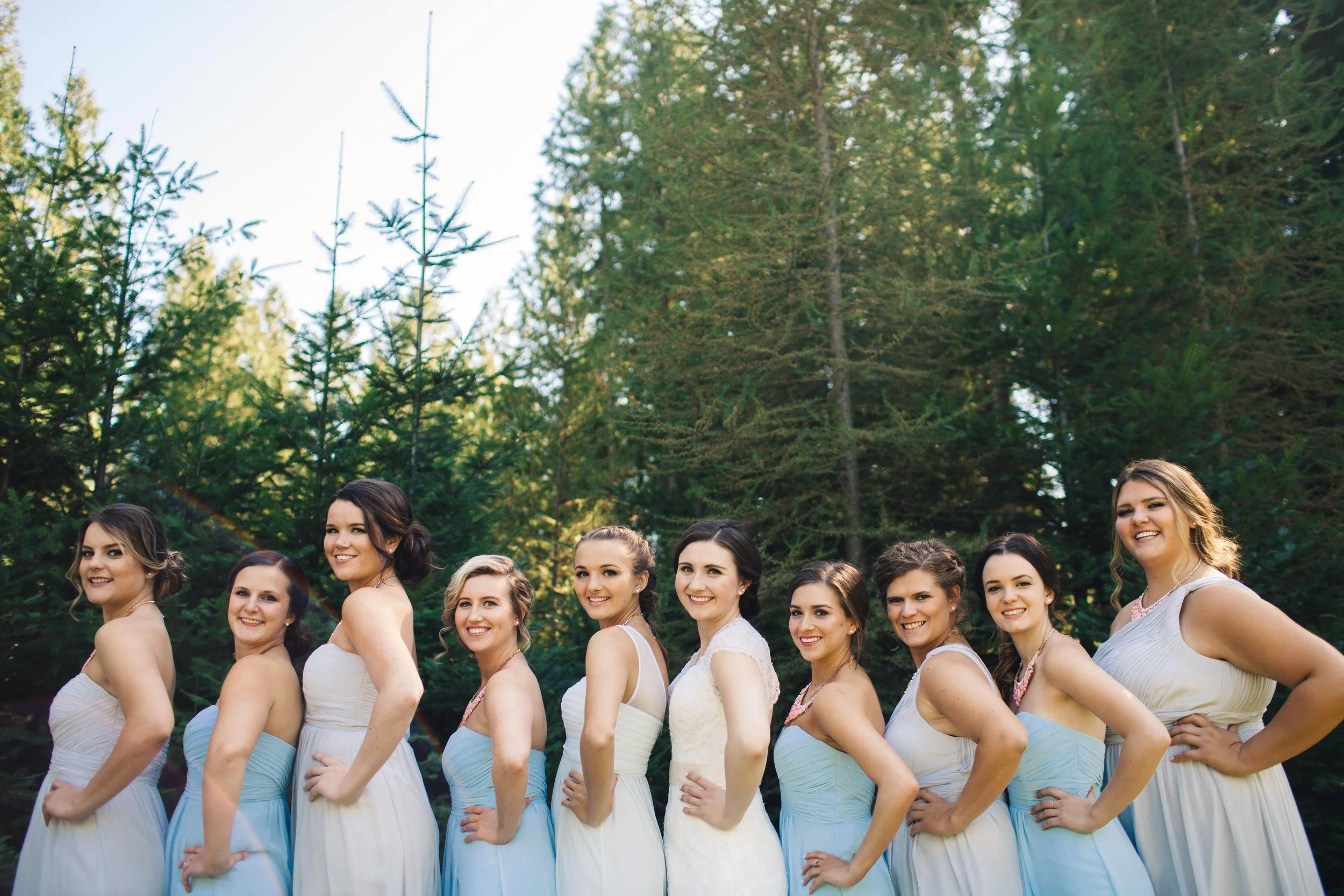 A group of ten women in formal dresses standing outdoors among pine trees, smiling and posing side by side.
