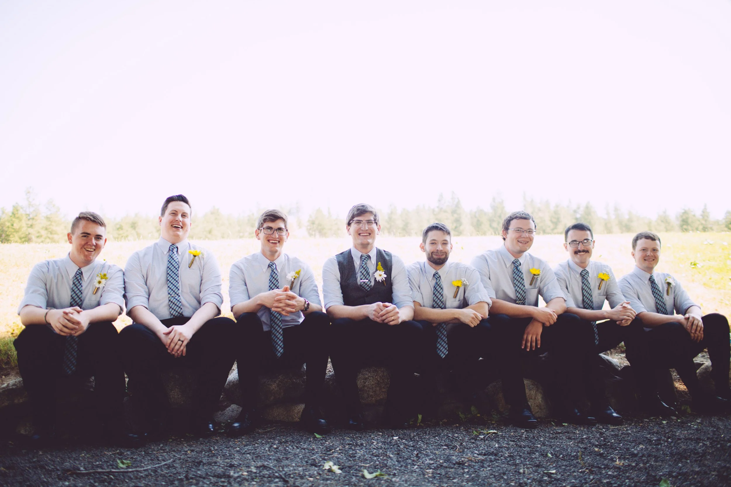 A group of nine men sitting on a stone wall outdoors during daytime, dressed in white shirts, black pants, and matching plaid ties, with yellow flowers pinned to their shirts, smiling and posing for a photo with a grassy field and trees in the backgr