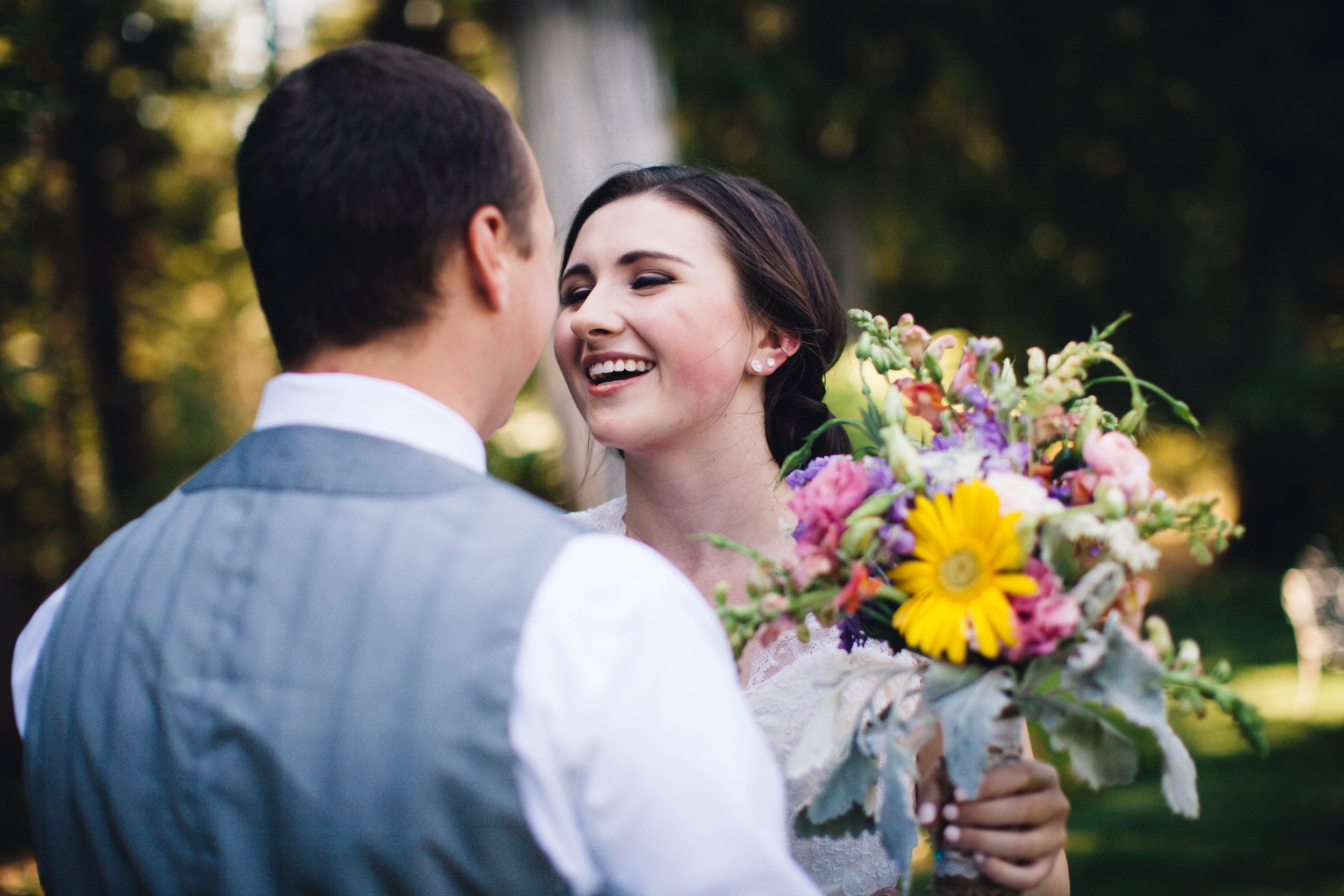 A woman holding a colorful bouquet of flowers smiling at a man with a gray vest and white shirt outdoors with trees in the background.