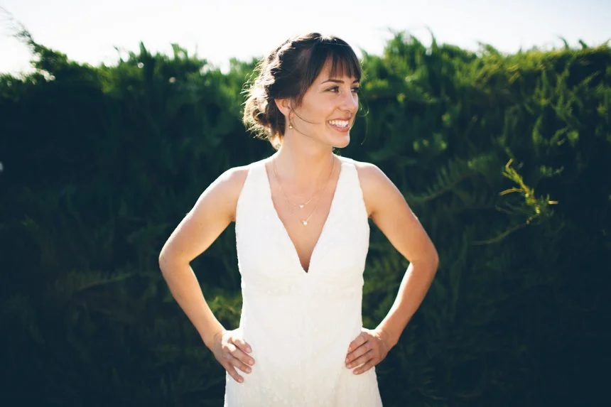 A woman with dark hair in a ponytail, wearing a white dress, smiling outdoors with greenery in the background.