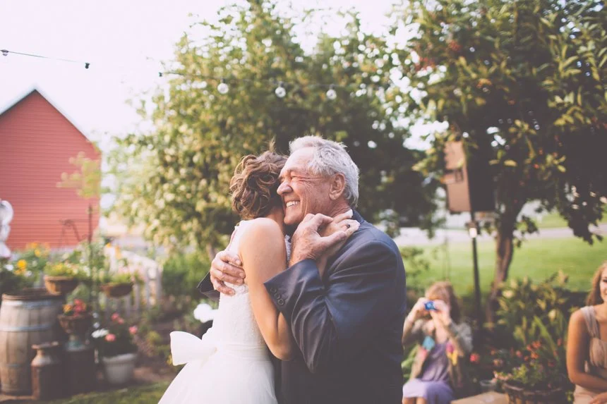 A woman and an elderly man hug and smile during an outdoor event, with trees, a red building, and other people in the background.
