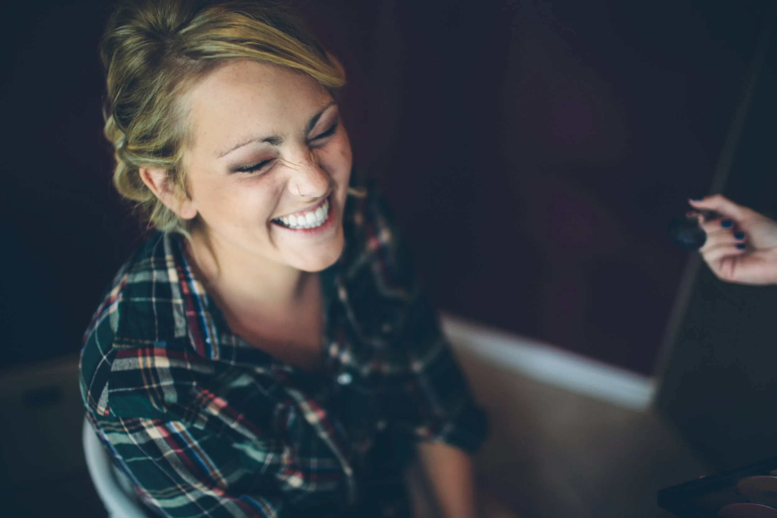 A young woman with short blonde hair and a nose piercing laughs with her eyes closed while sitting in a studio. A person's hand holding a camera is visible on the right side of the image.