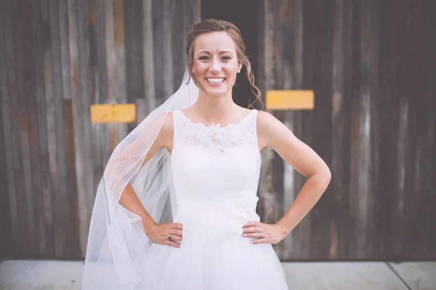 A woman in a wedding dress smiling with hands on hips in front of a wooden barn door.