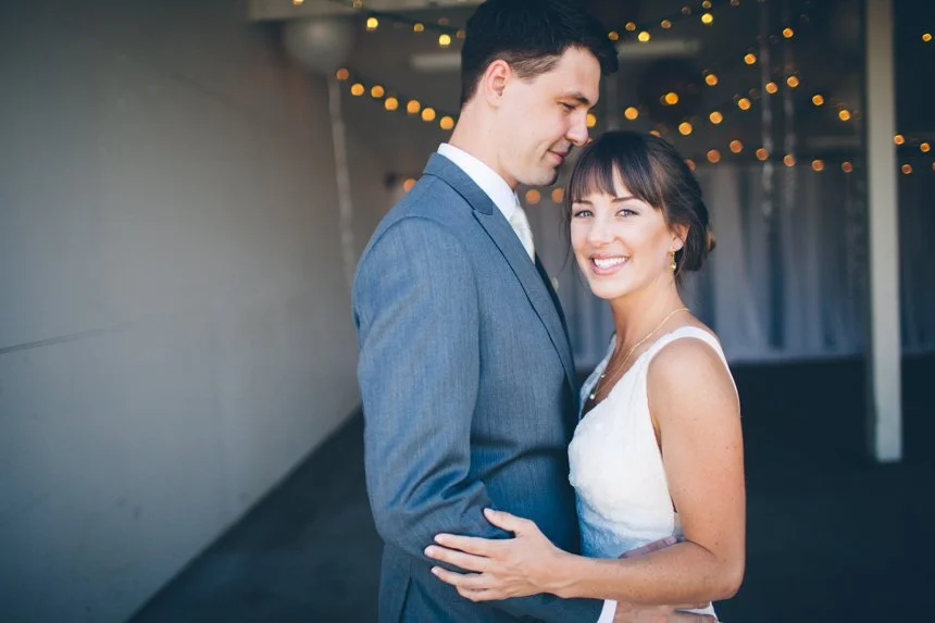 A smiling couple dressed in formal attire, with the woman in a white dress and the man in a blue suit, embracing each other at a wedding or special event, with string lights in the background.