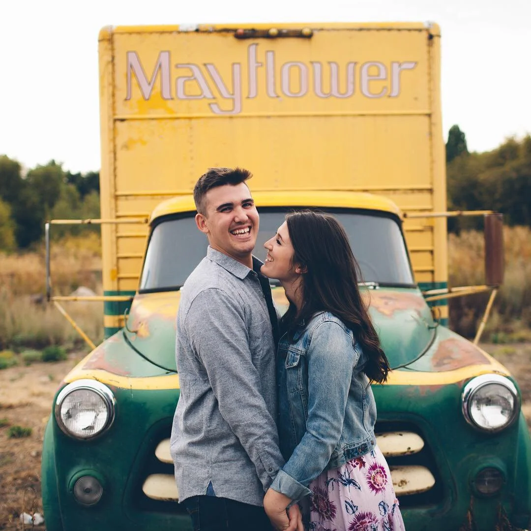 A smiling couple standing in front of a colorful Mayflower truck, holding hands and looking into each other's eyes.