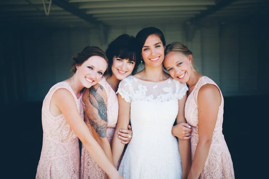 Four women in lace dresses smiling and hugging each other at a wedding or special event.