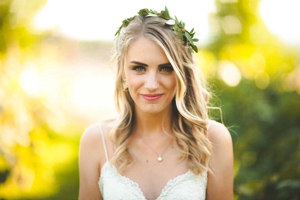 A woman with long blonde hair wearing a white dress and a green leaf crown, stands outdoors with a background of blurred greenery and sunlight.