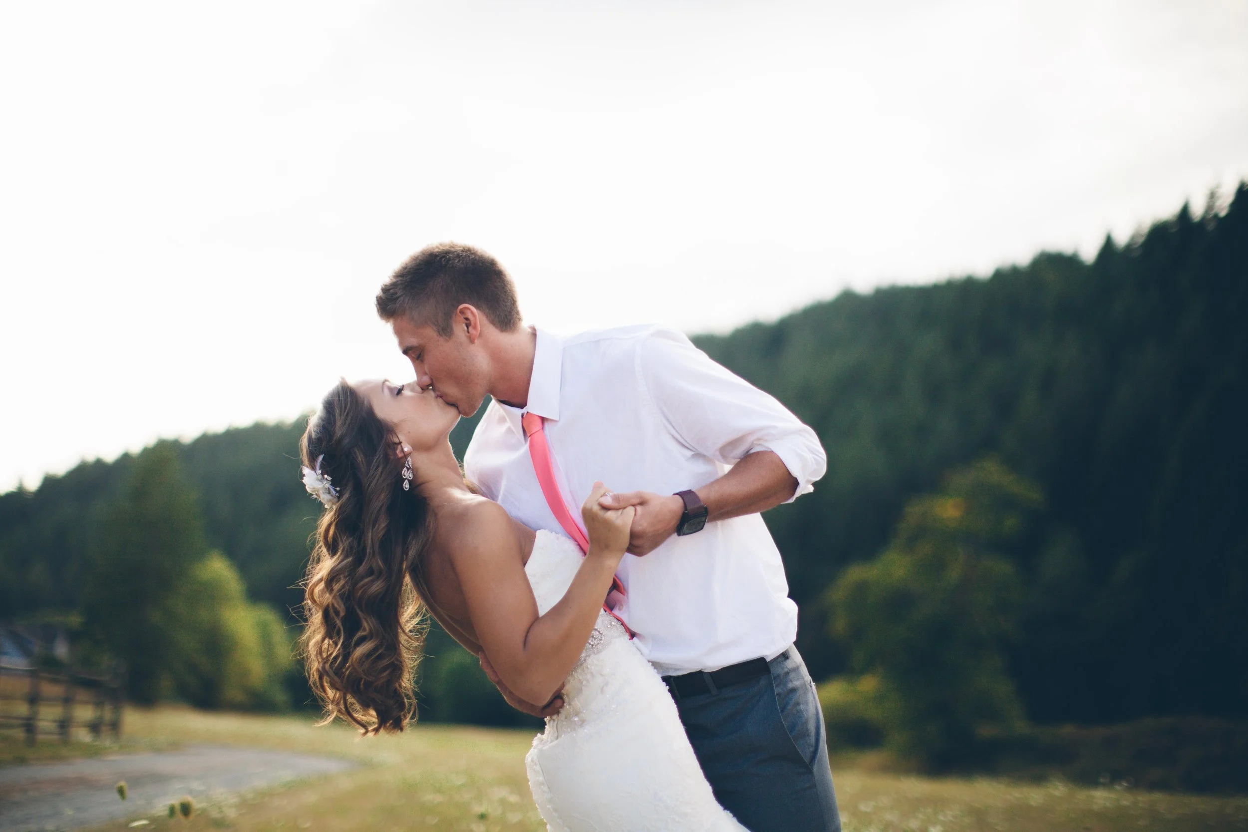 A newlywed couple sharing a kiss outdoors, with the groom in a white shirt and pink tie, and the bride in a wedding gown with long, wavy hair, surrounded by a scenic landscape of trees and open sky.