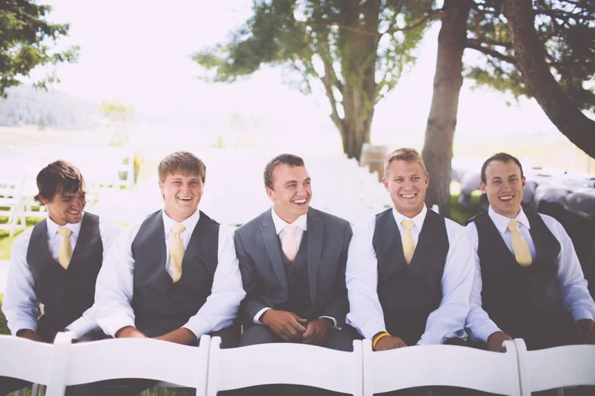 Group of five men in suits and vests sitting outdoors, smiling, under trees, at a formal event.