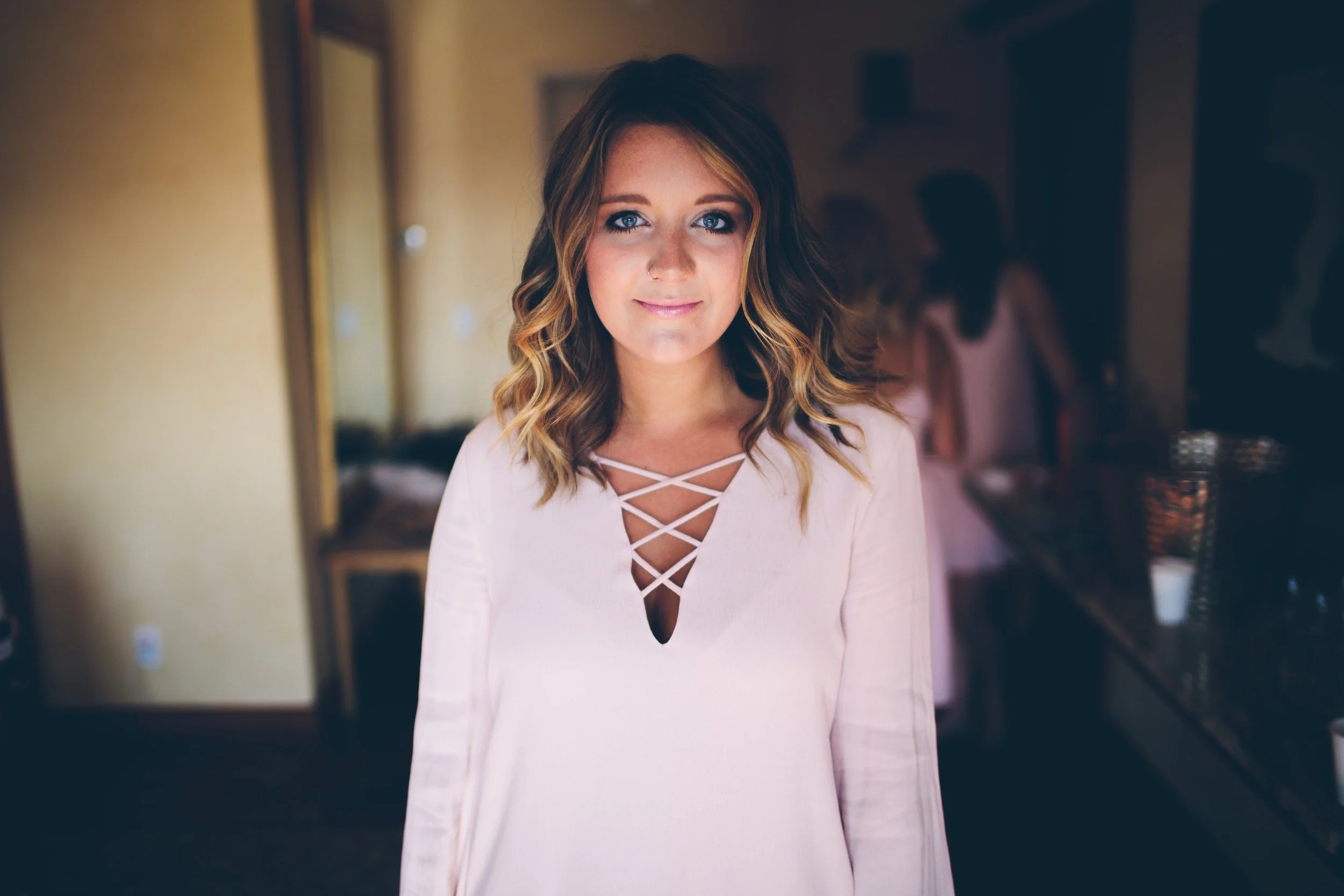 Young woman with wavy light brown hair and blue eyes, wearing a light pink dress with crisscross neckline, standing indoors in a softly lit room with other women in the background.