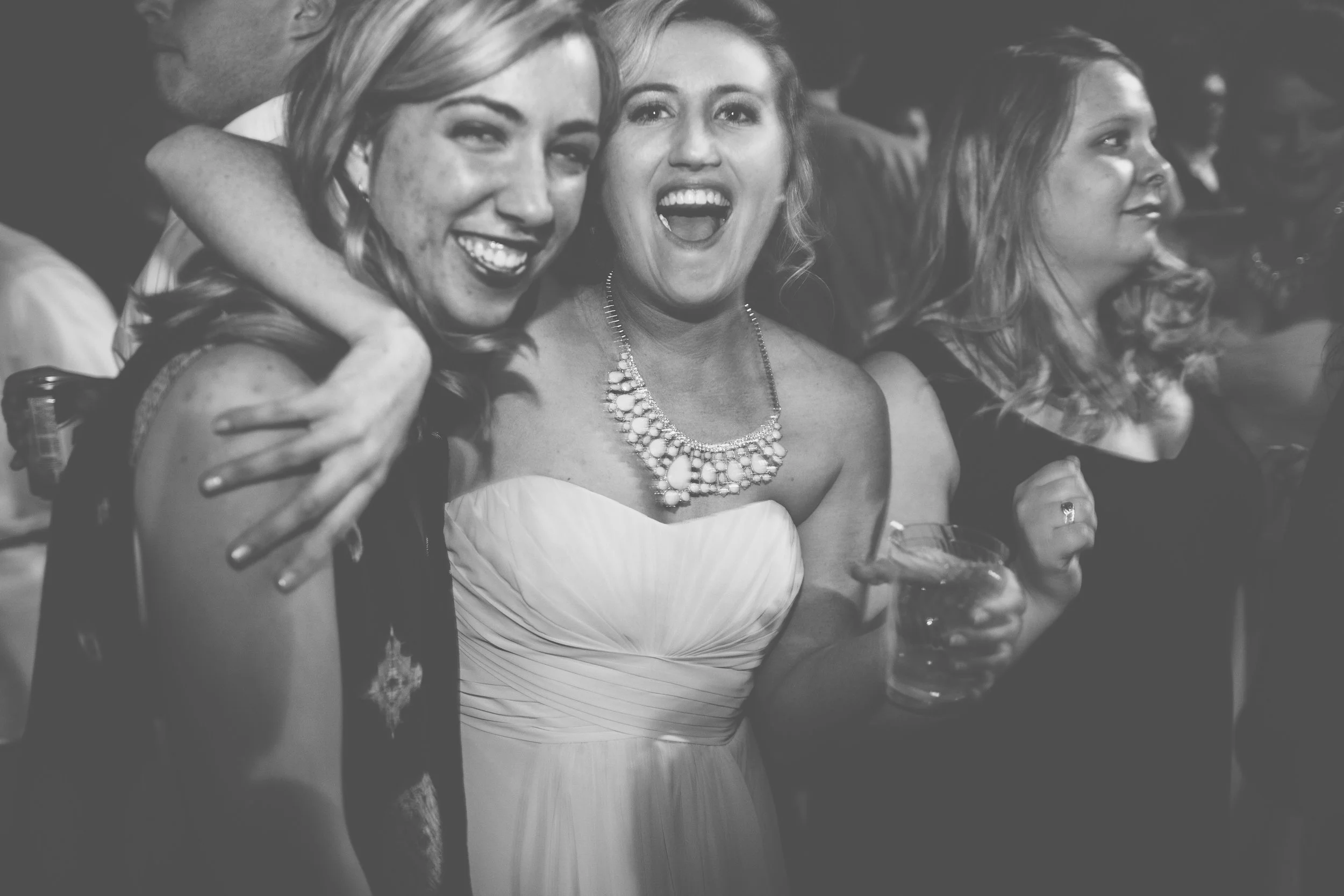Group of women at a celebration, smiling and holding drinks, with one woman in the center wearing a strapless dress and a large pearl necklace.