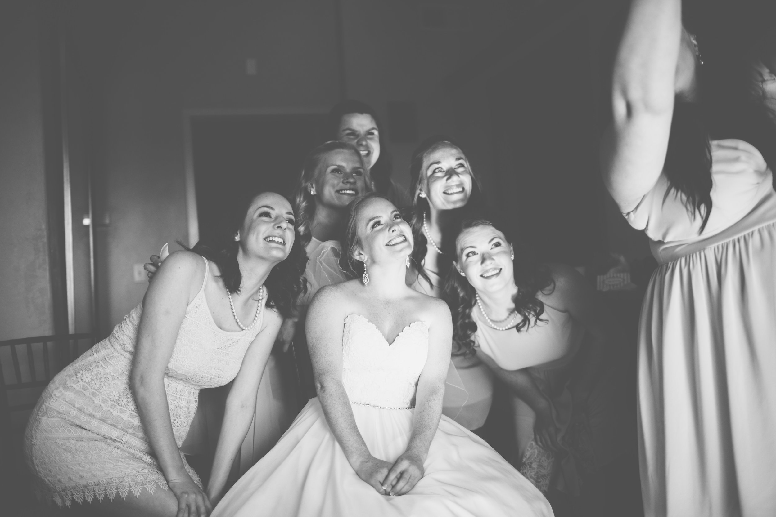 Group of women, including a bride in a wedding dress, smiling and looking at a woman in a dress, in a bridal or wedding setting.