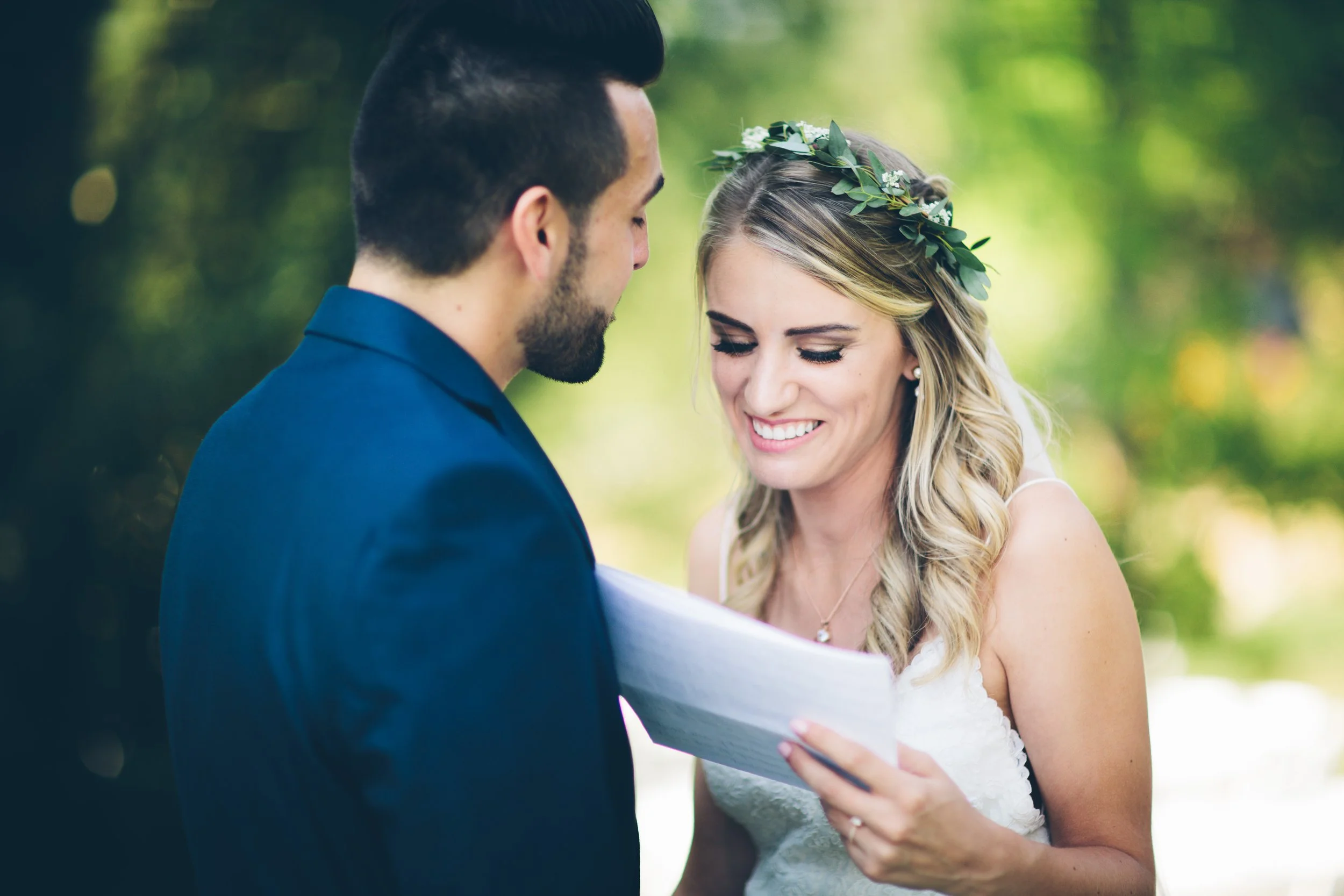 A bride and groom during their wedding ceremony outdoors, with the bride smiling and reading vows from a paper, wearing a white dress and a floral crown, and the groom dressed in a blue suit, set against a blurred green natural background.