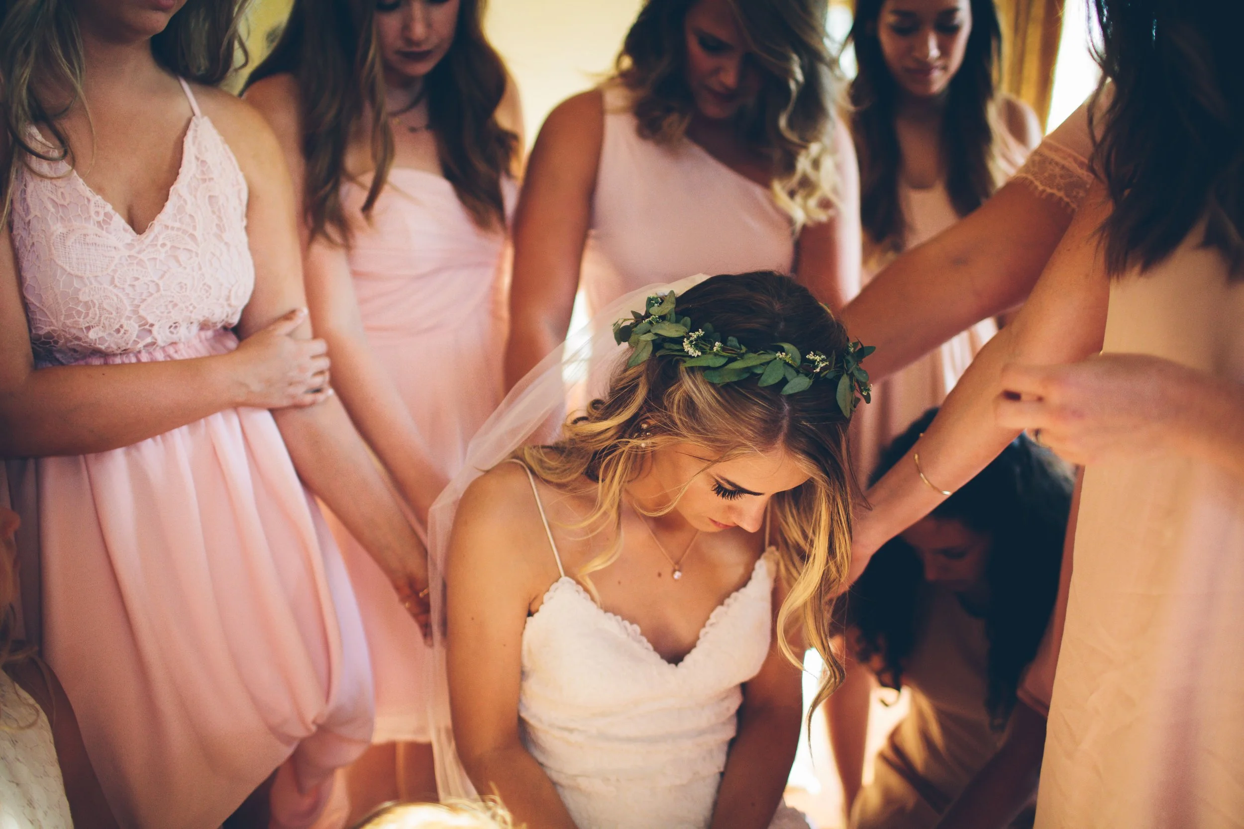 Bride with long blonde hair, wearing a wedding dress and a green floral crown, is bowed down with other women around her, who are dressed in pink and peach dresses, participating in a spiritual or prayerful moment.