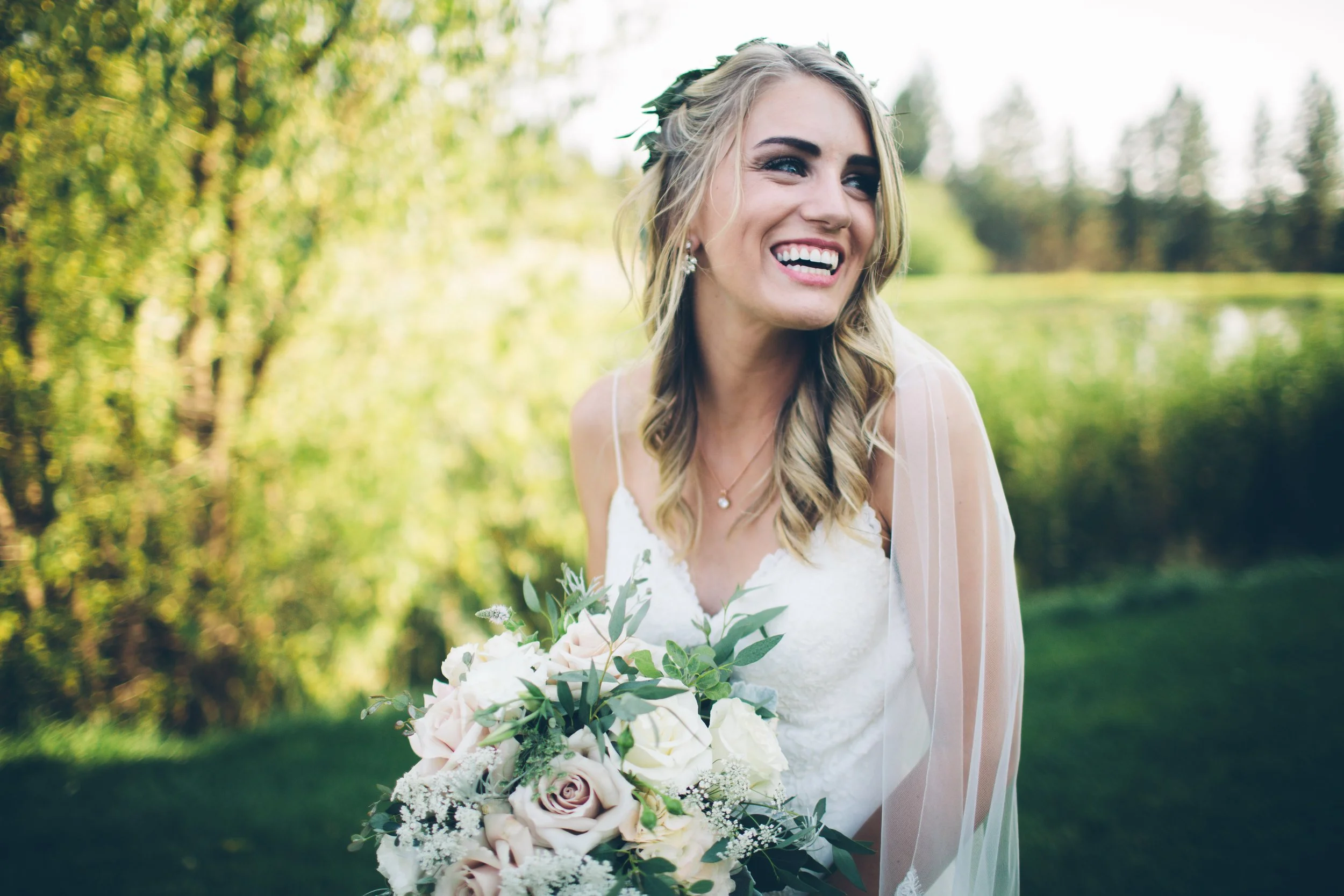 A smiling bride in a white wedding dress holding a bouquet of roses and greenery outdoors with trees and grass in the background.