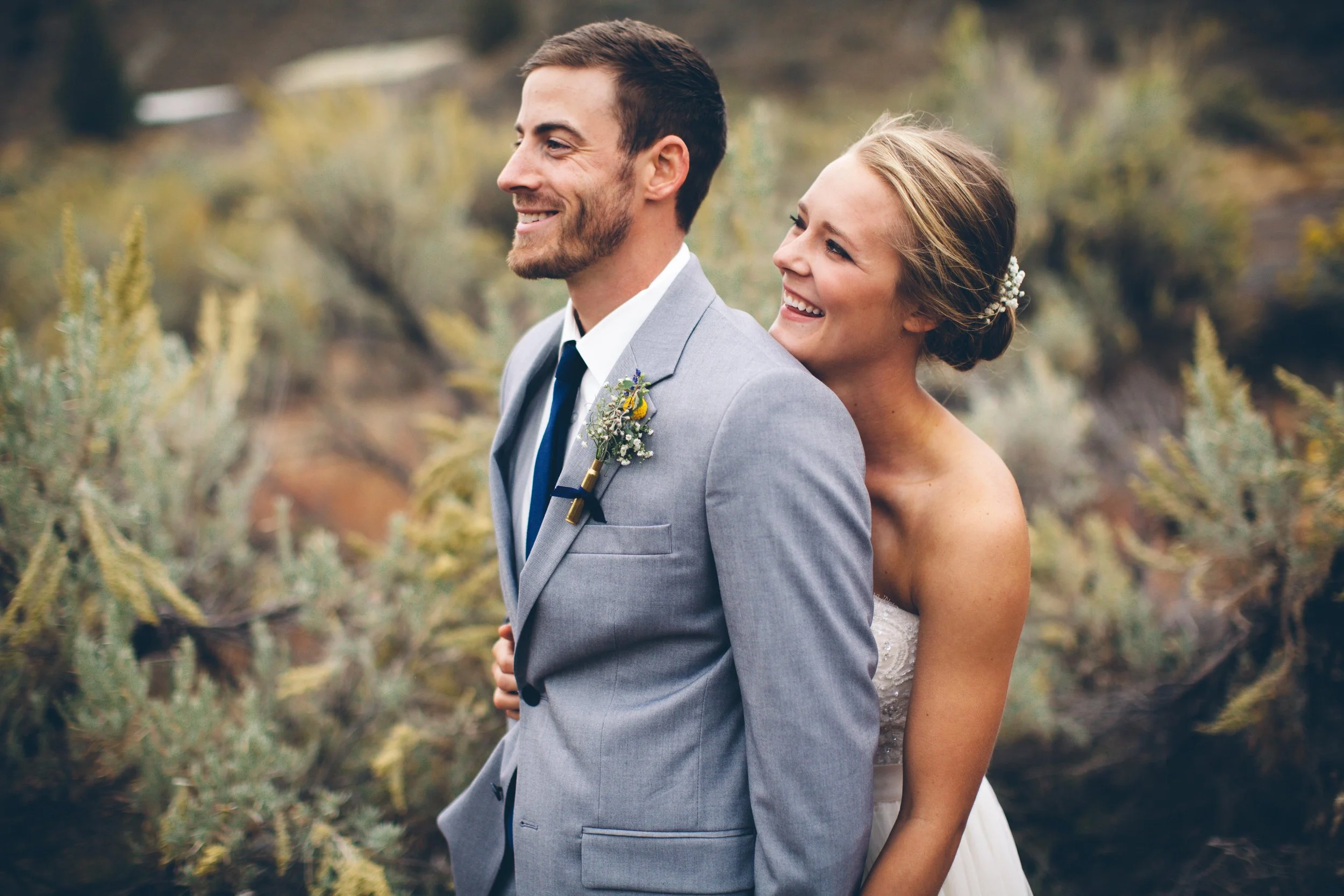 A bride and groom in wedding attire outside, with the bride embracing the groom from behind, both smiling, natural background of shrubs and trees.