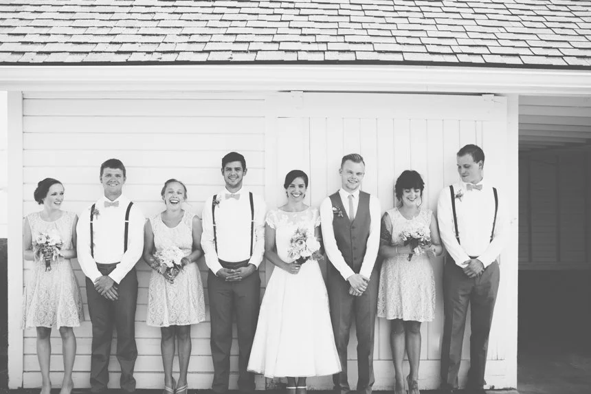 A group of wedding party members, including the bride and groom, standing in front of a white building, all smiling and holding flowers.
