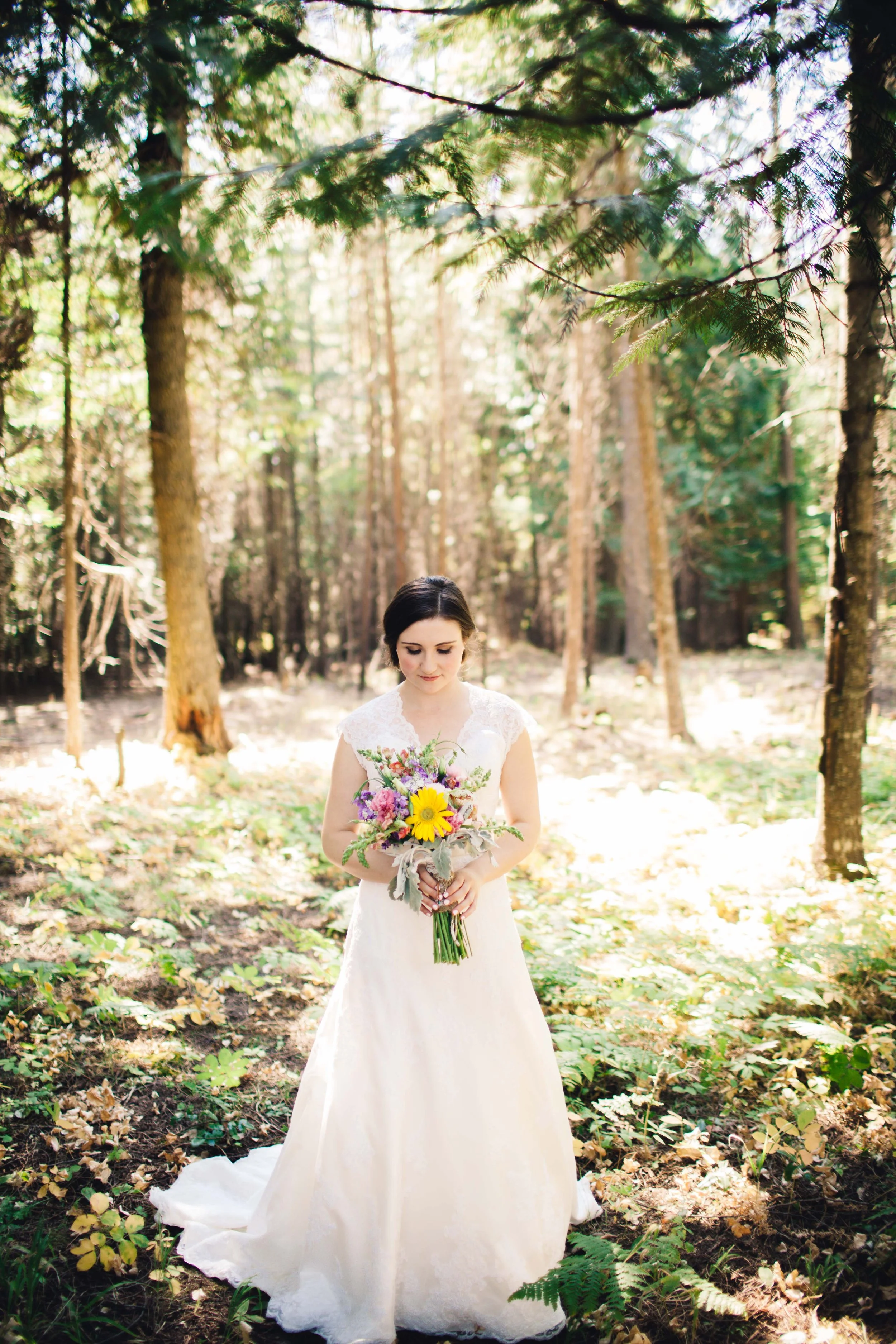 A bride in a white wedding dress holding a colorful bouquet of flowers, standing in a sunlit forest surrounded by tall trees and greenery.