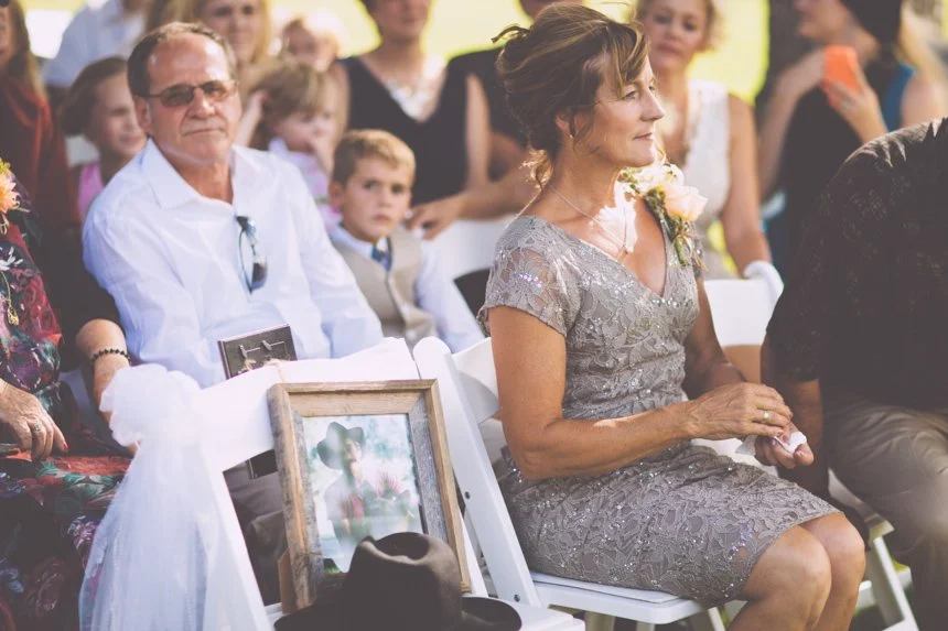 A woman in a silver dress sitting at an outdoor event, likely a wedding, with people gathered around her. There is a framed photo and some flowers in her lap.