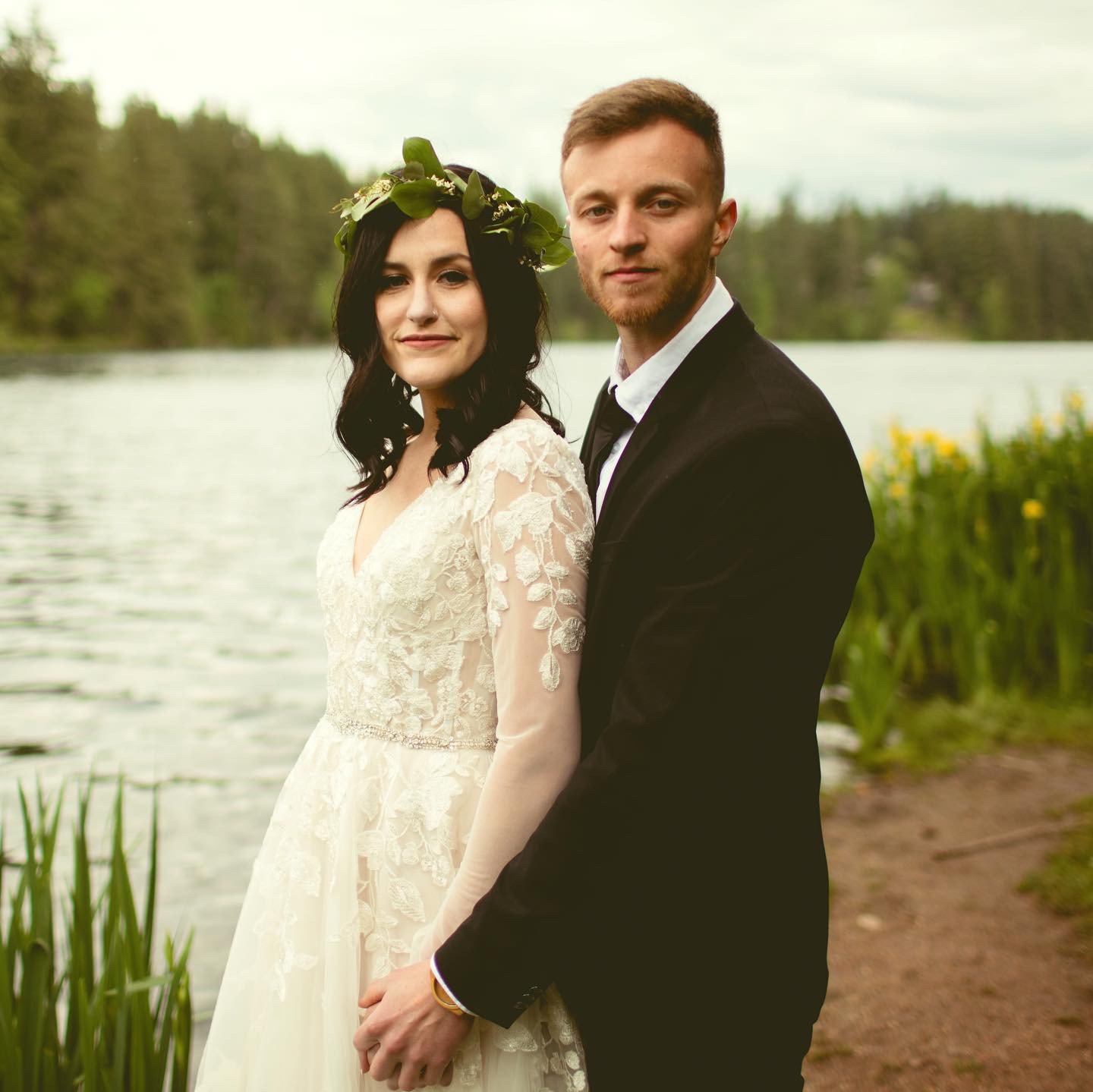 A bride in a white lace wedding dress with dark hair and a floral crown, standing close to a groom in a black suit, by a lake with trees in the background.