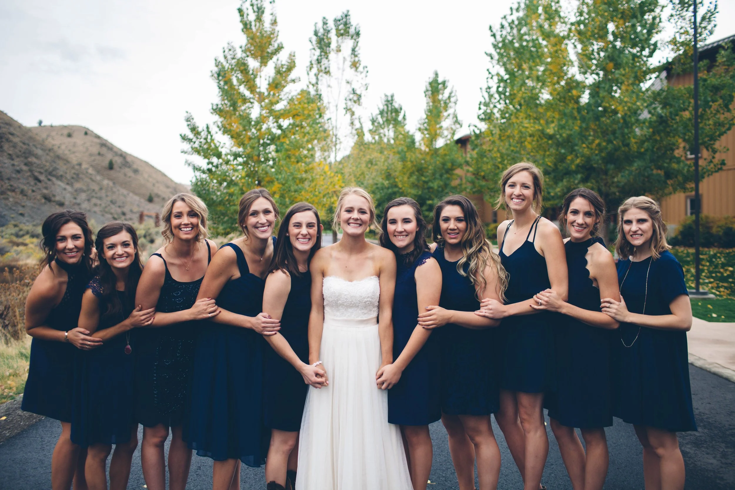 A bride in a white dress holding hands with a bridesmaid, surrounded by her bridesmaids in navy blue dresses, standing outdoors with trees and buildings in the background.