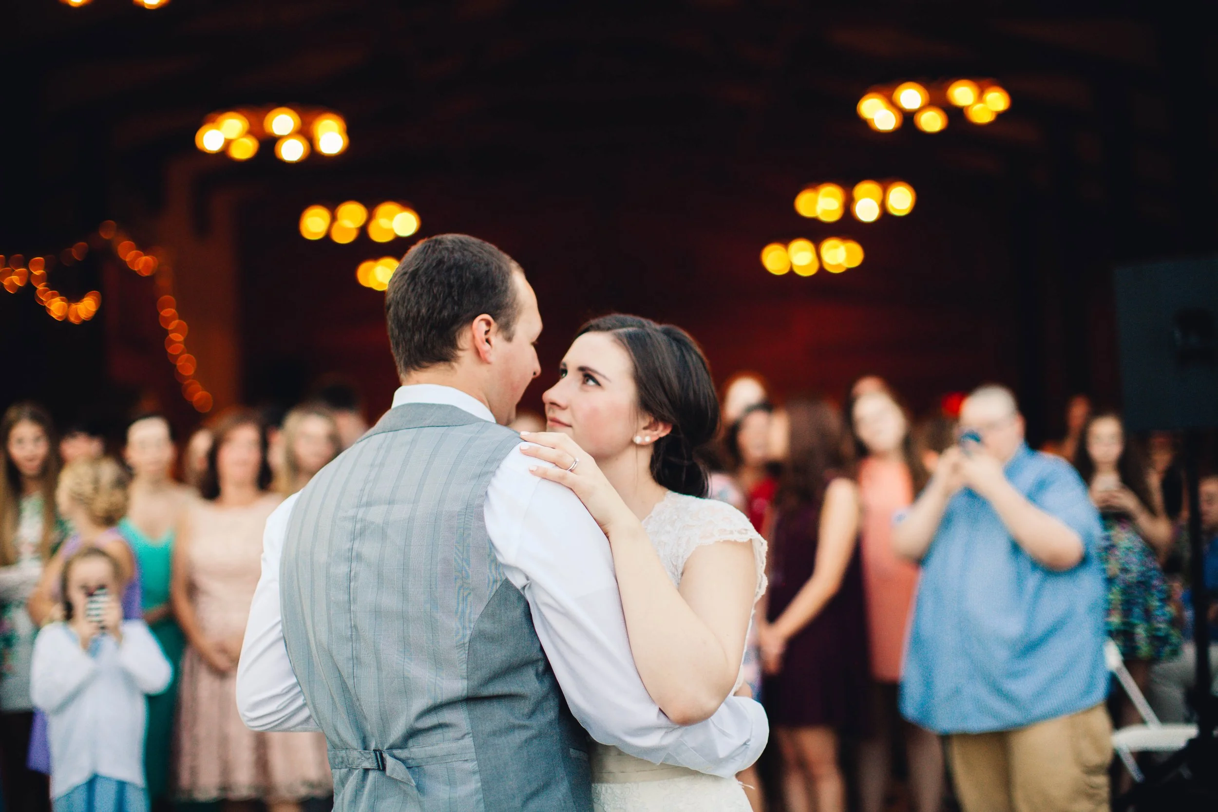 A bride and groom sharing a dance during their wedding reception, surrounded by guests in a warmly lit venue.