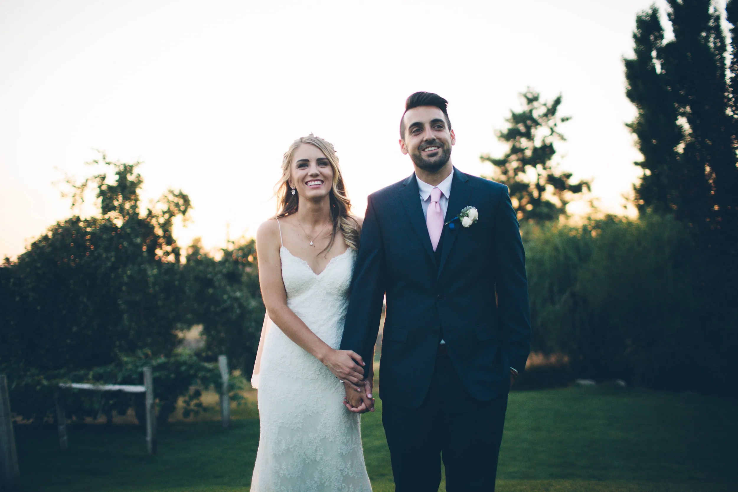 A newlywed couple holding hands outdoors during sunset, the bride in a white lace wedding dress and the groom in a dark suit with a pink tie, both smiling.