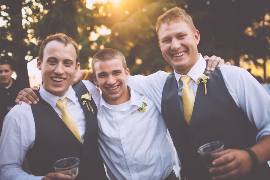 Three men smiling and standing close together outdoors during sunset, two wearing vests and ties, one in a white shirt, with trees in the background.